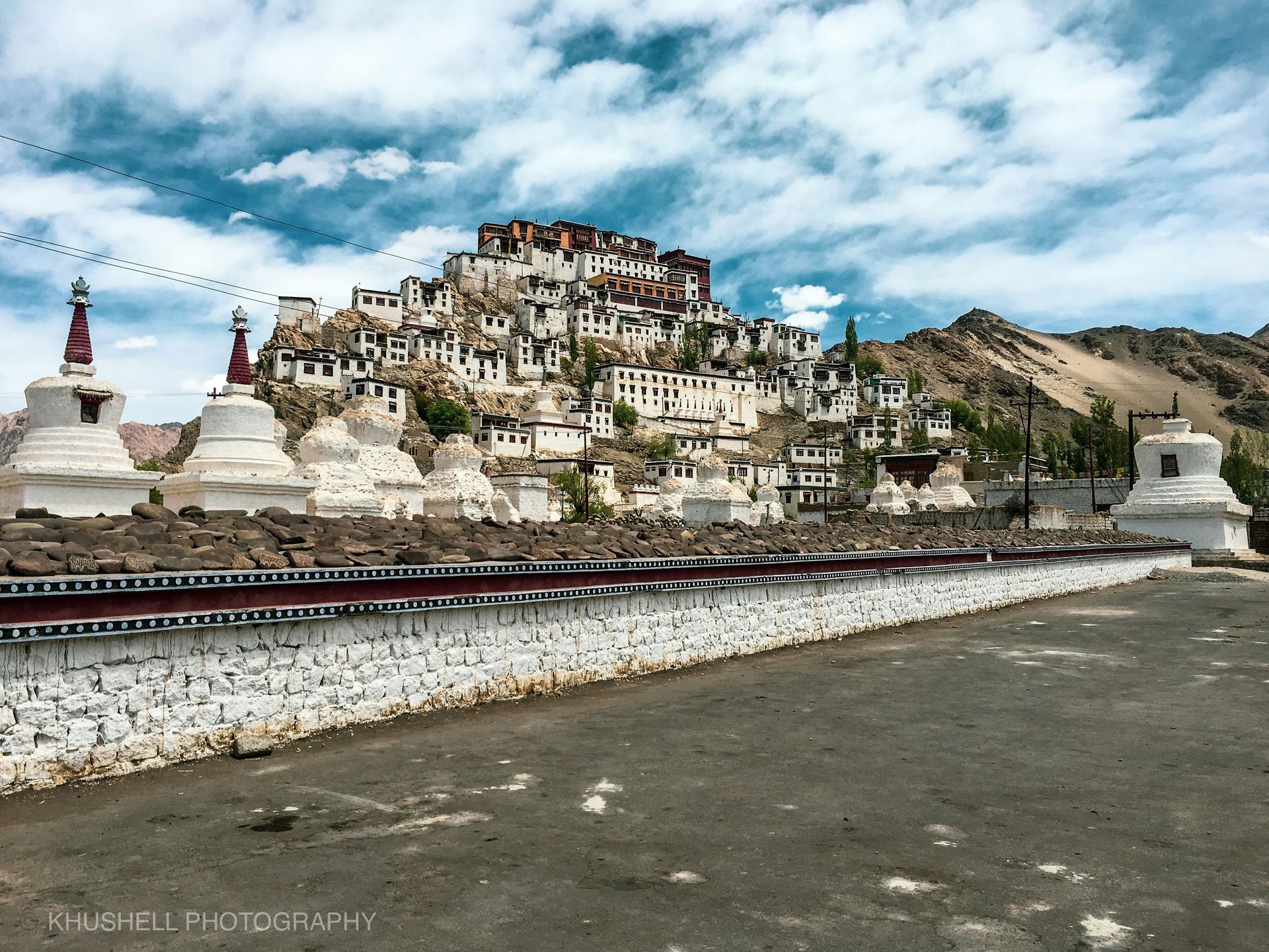 White monastery perched on a hillside in Ladakh, India, with prayer flags and stupas under a cloudy sky.