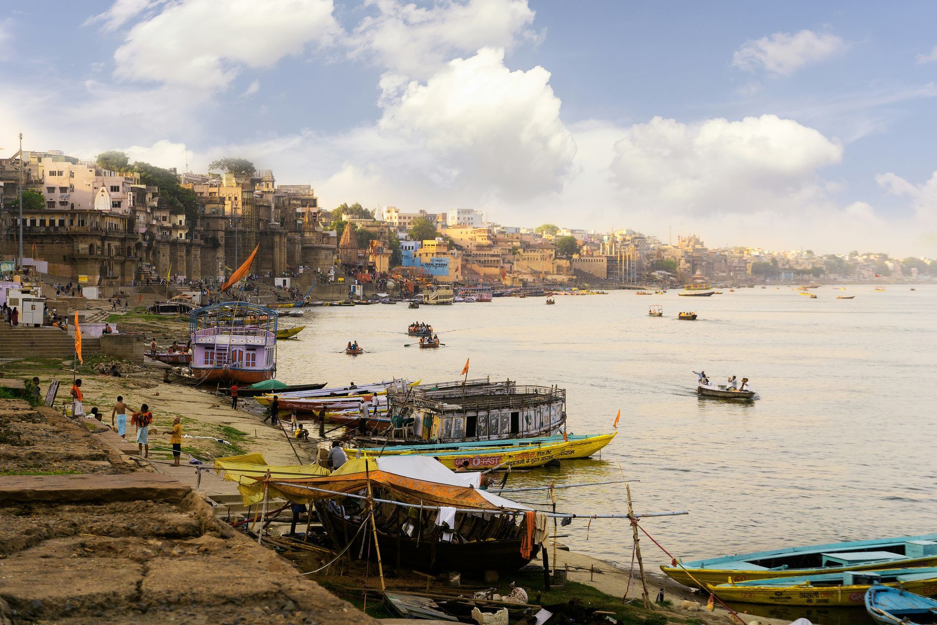 Ghat along Ganges River in Varanasi, India, with boats, buildings, and people on the shore and in the water.
