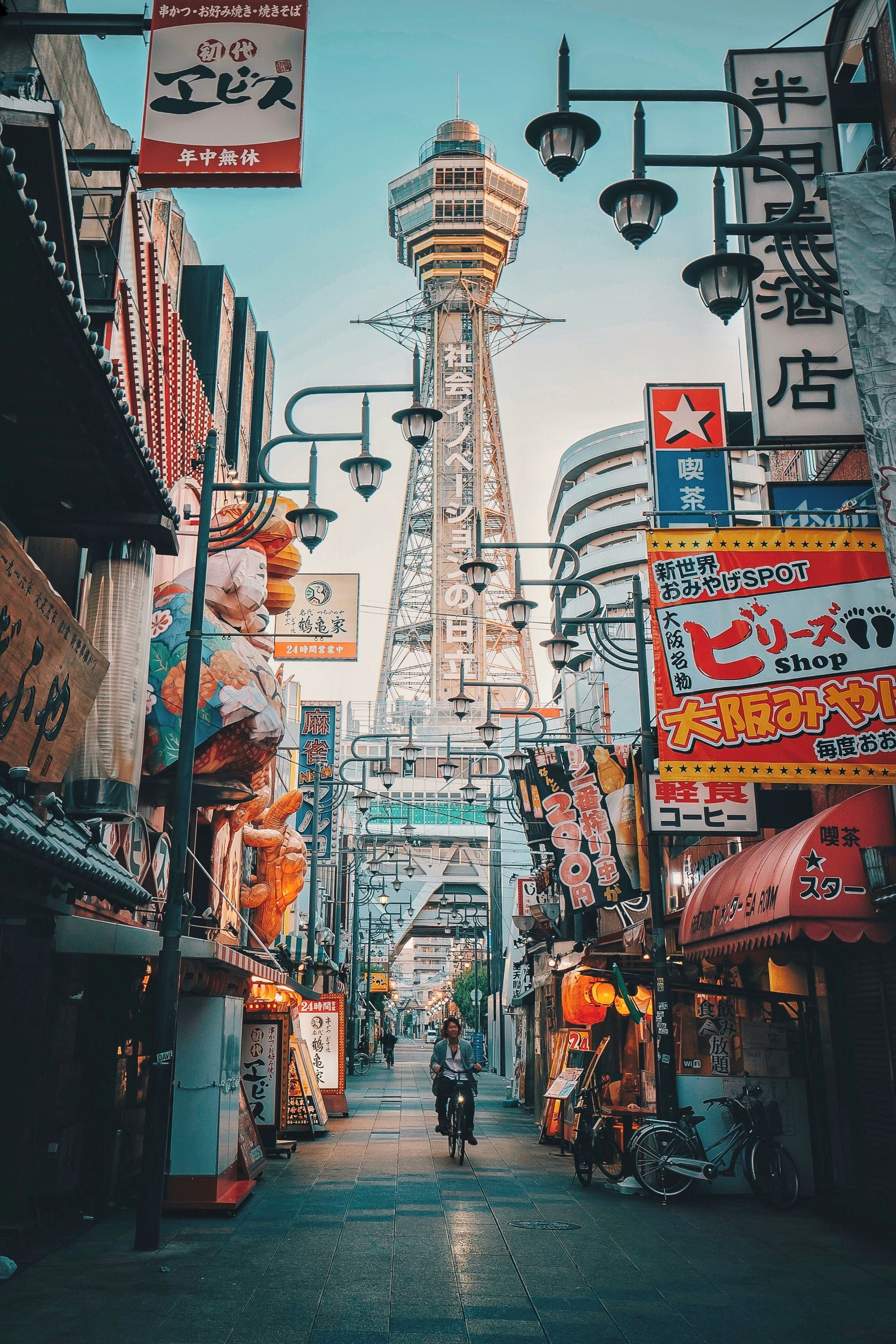 Busy street scene in Japan with Tsutenkaku Tower. Buildings with neon signs line the street; one person walks toward the tower.