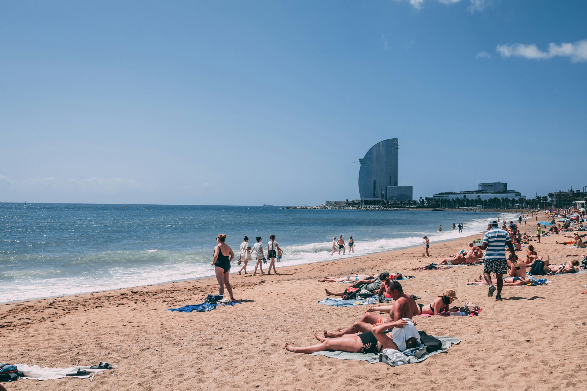 People sunbathing and walking on Barceloneta beach, with the W Barcelona hotel in the background.