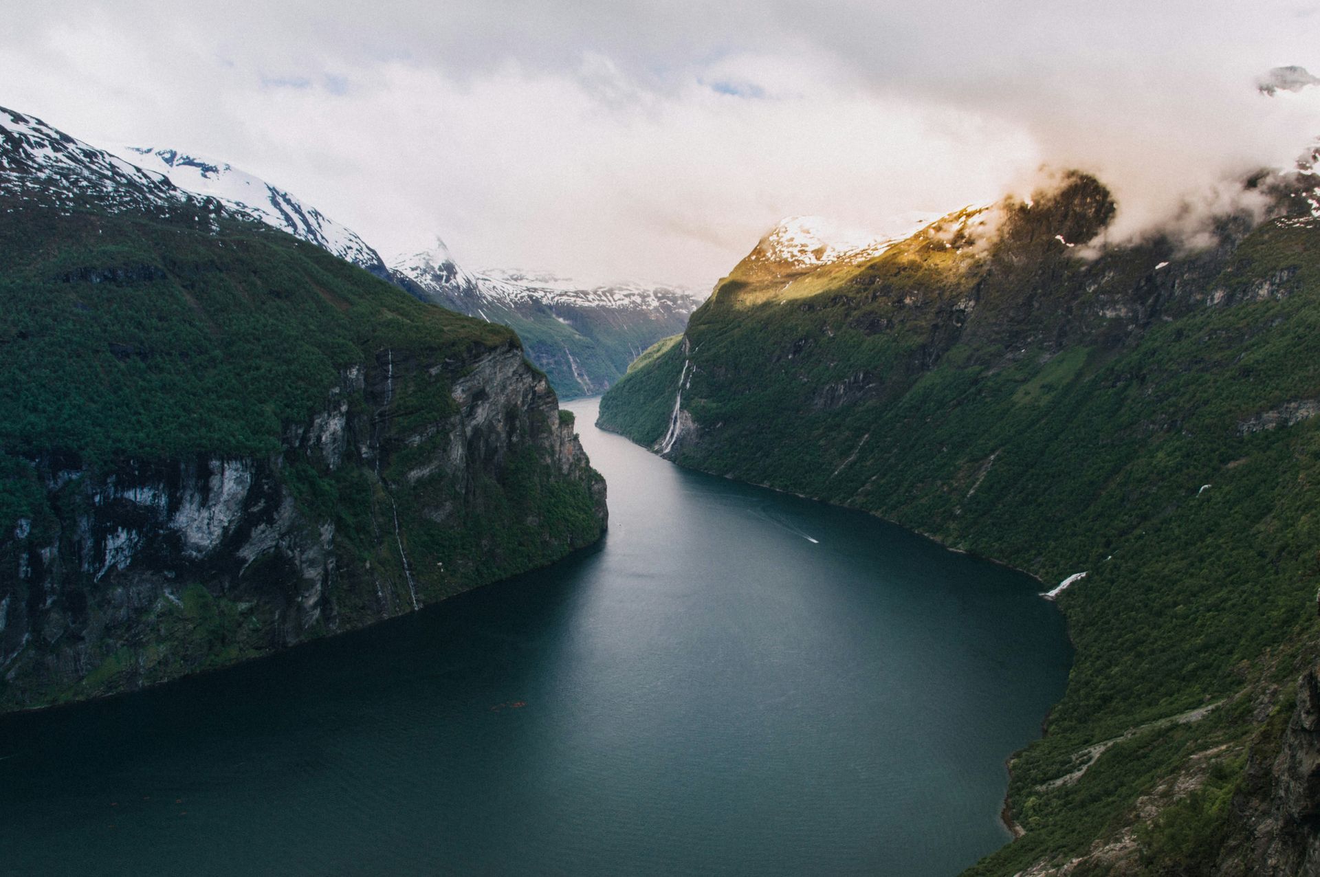 The Geirangerfjord between towering green cliffs, snow-capped peaks and overcast skies in Norway.