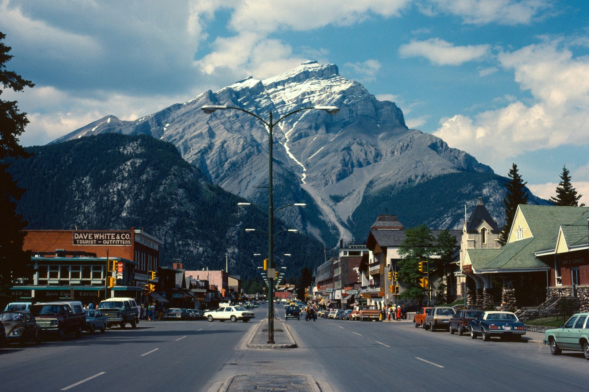 Street scene in Banff, Canada, with buildings lining the road and a large, snow-capped mountain in the background under a partly cloudy sky.