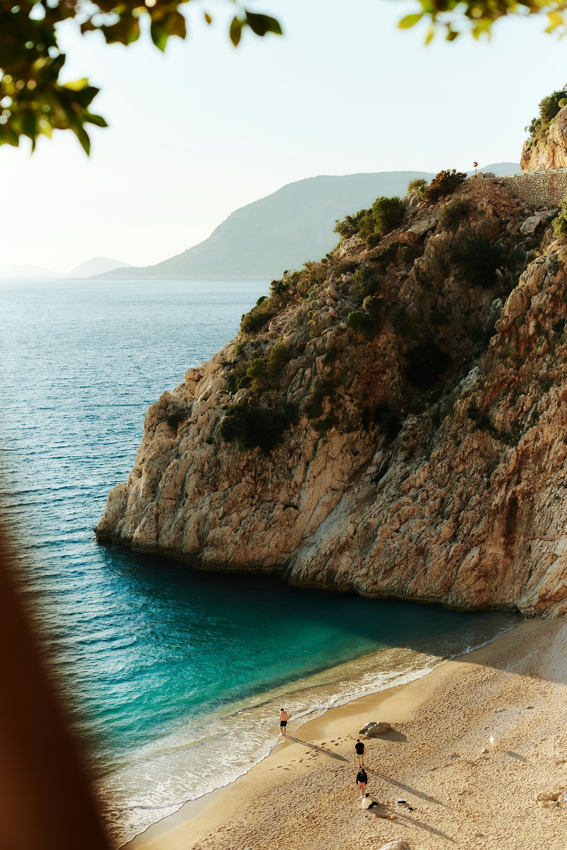Rocky cliffside meets turquoise water; beach below with people walking in Kaş, Turkey.