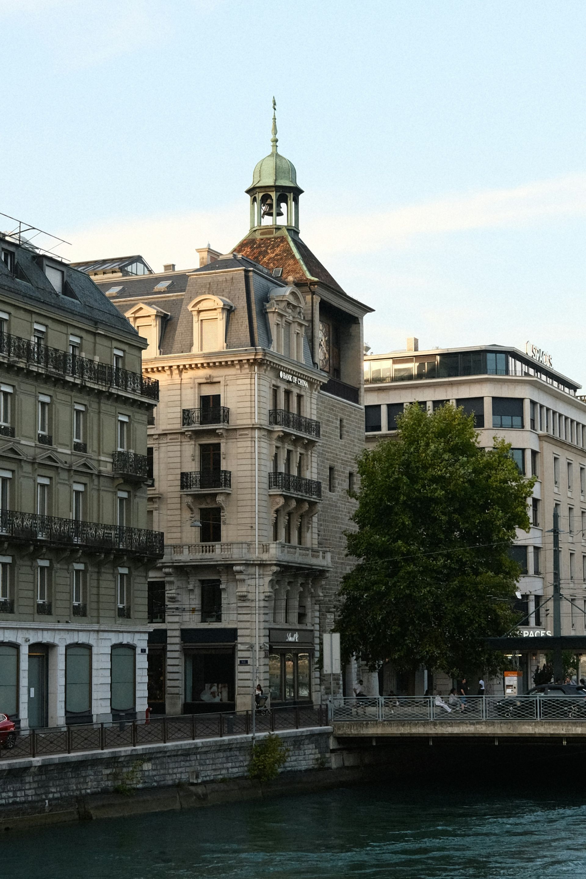 Buildings along a waterway in Geneva, Switzerland. A tree and a bridge are also visible.