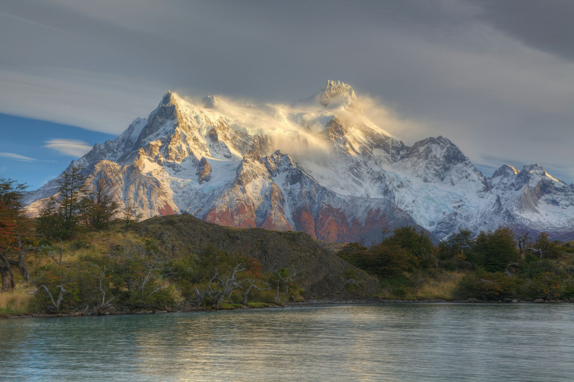 Snow-capped mountain illuminated by sunlight, overlooking a calm lake in Torres del Paine, Chile.