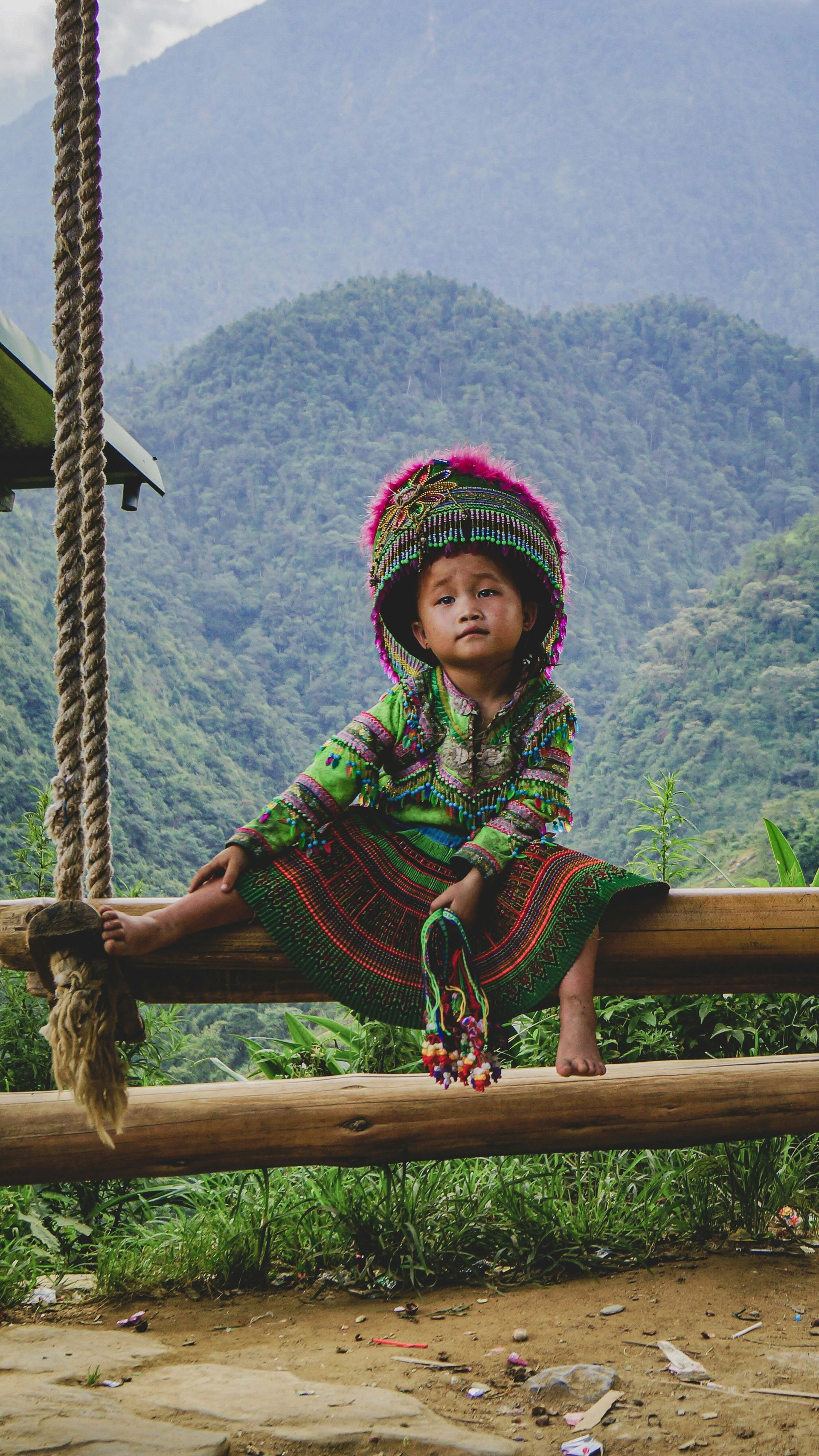 Young girl in traditional dress sits on a wooden swing, mountain backdrop in Sapa, Vietnam. 