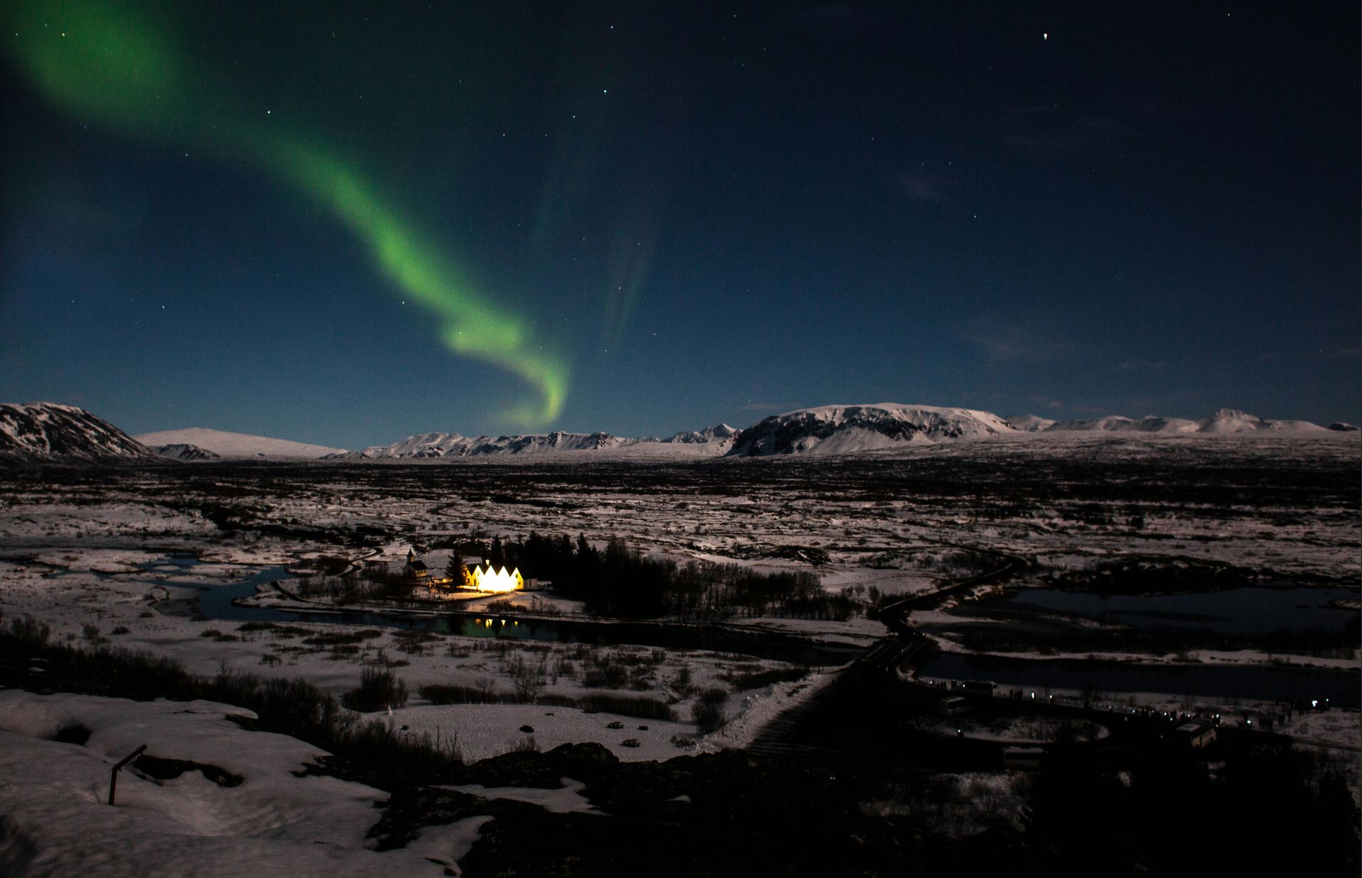 Northern Lights over snow-covered landscape, illuminated building, mountains in the distance on the Golden Circle, Iceland.