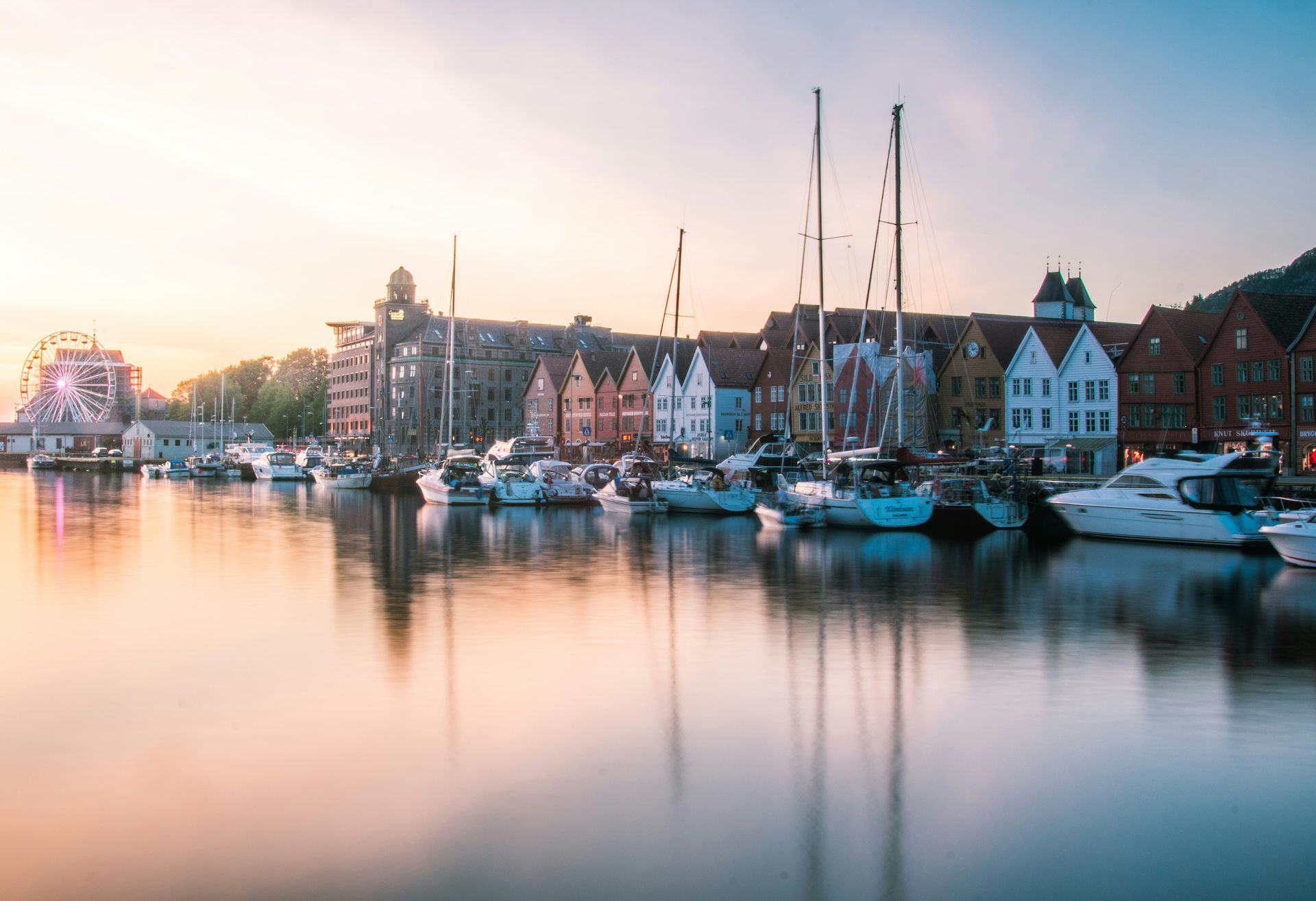 Harbor with boats docked, colorful buildings, and Ferris wheel under a pastel sunrise in Bergen, Norway.
