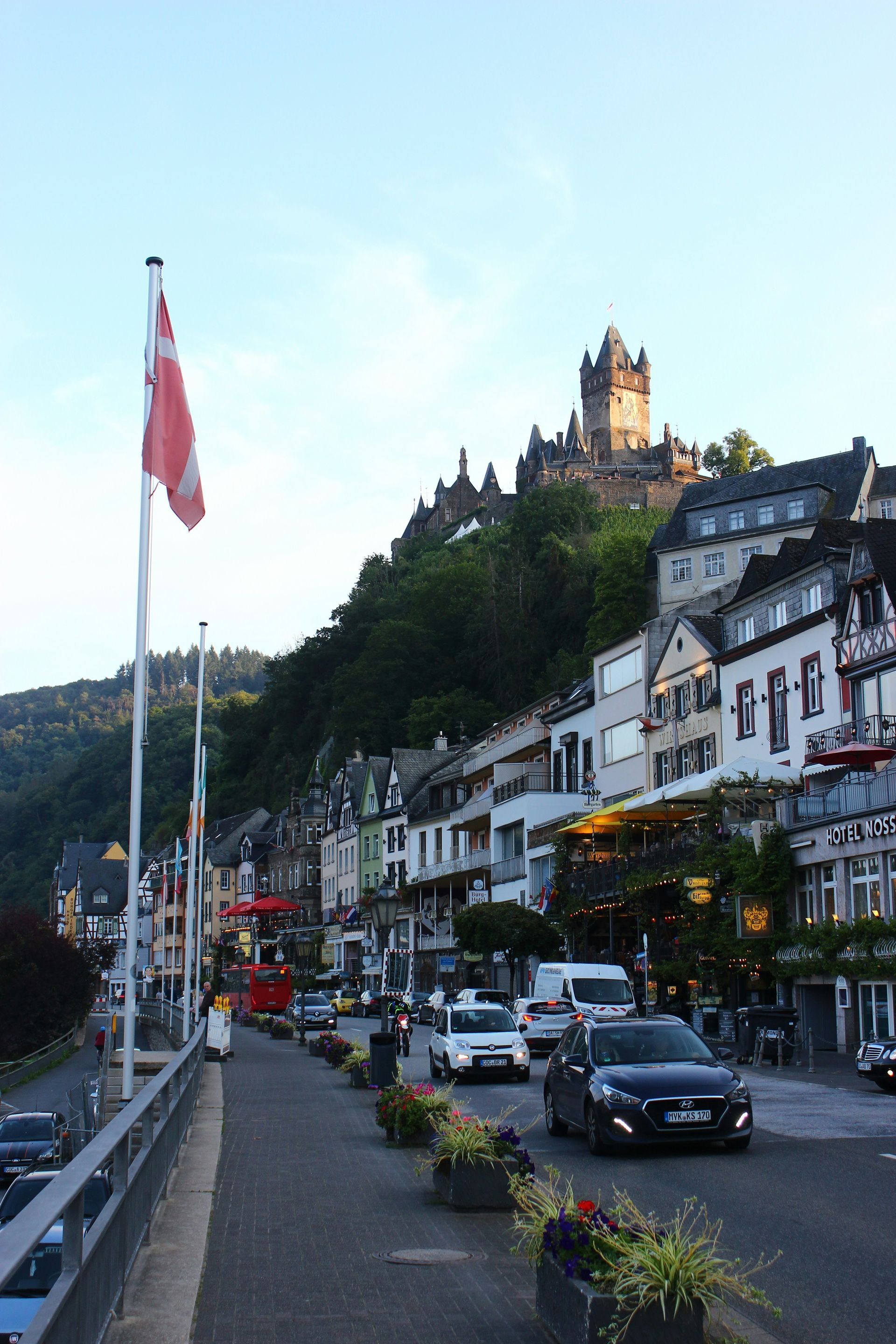 Roadside view in Cochem, Germany, with castle atop hill, cars, buildings, and flags.