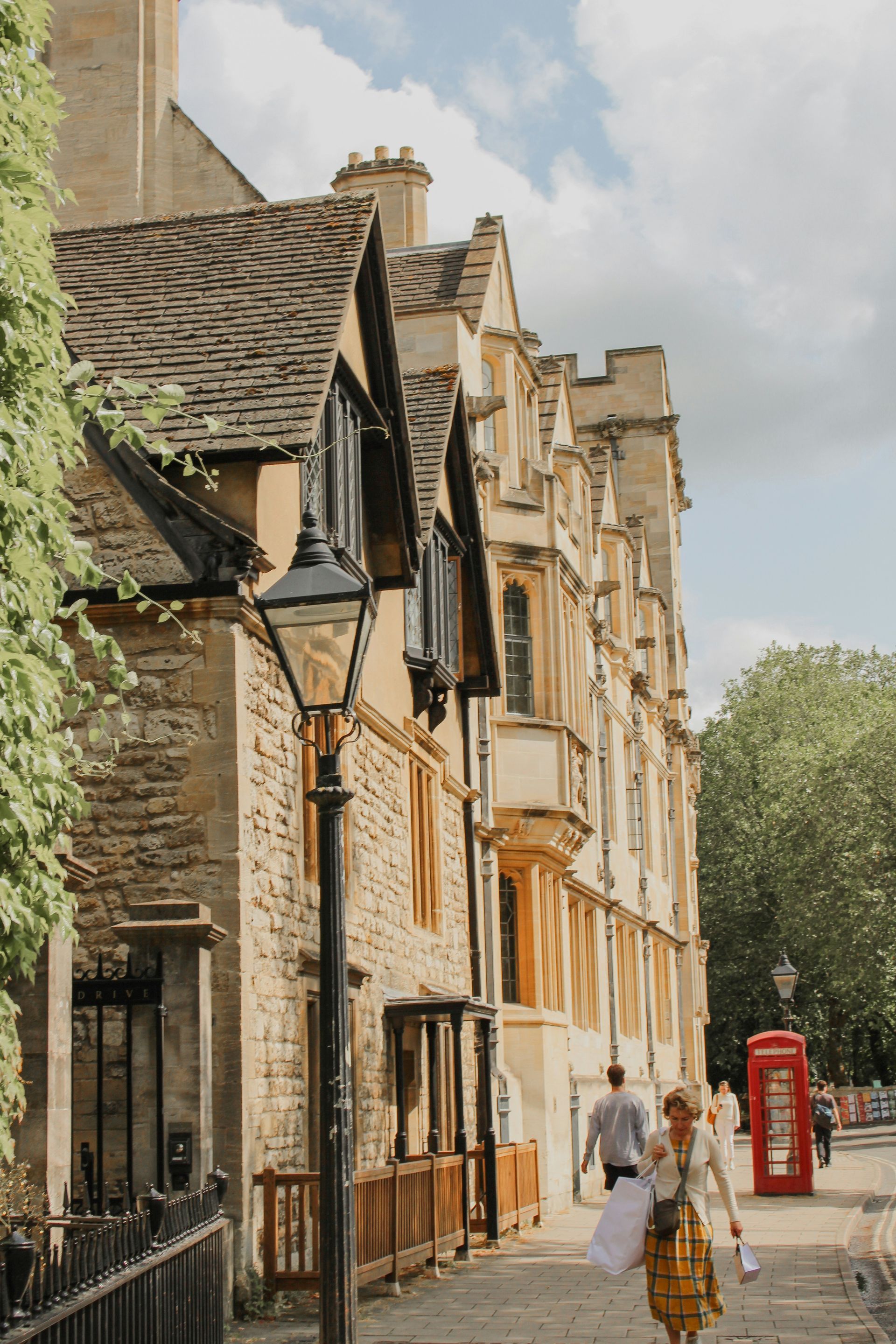 Old stone buildings and a red phone booth line a sidewalk with people walking on a sunny day in Oxford, England.