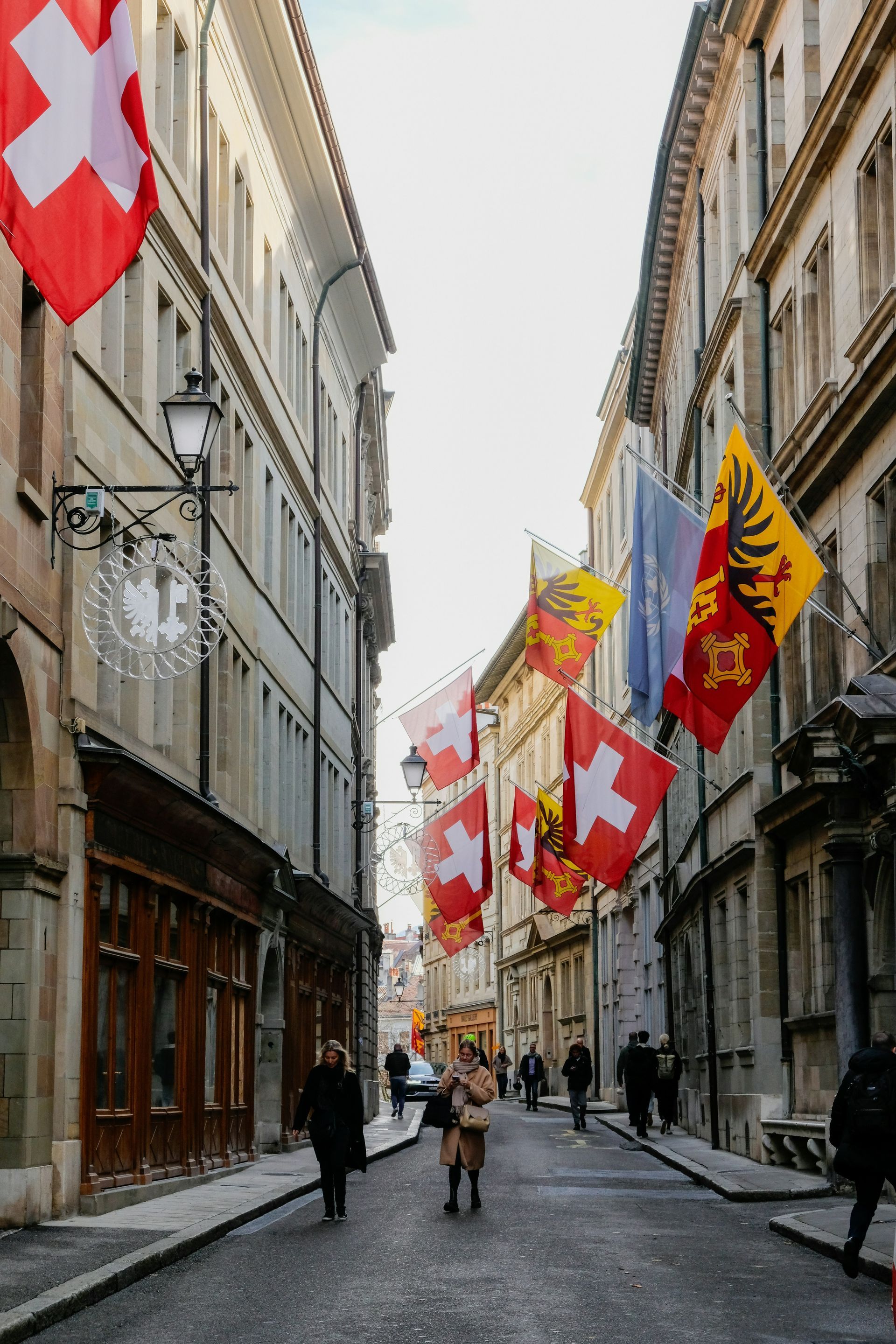 Narrow street, lined with old buildings and Swiss flags, pedestrians walking in Geneva, Switzerland.