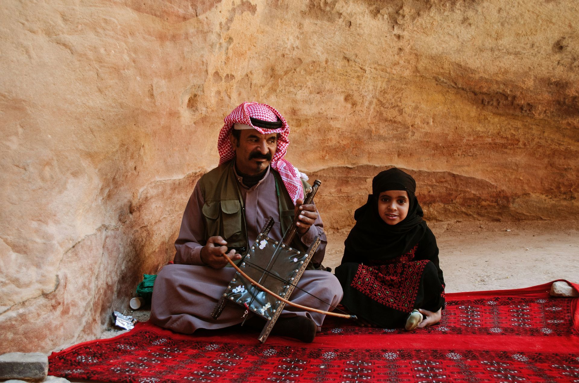 Man playing a stringed instrument, seated next to a child, on a red rug in a stone setting in Jordan.