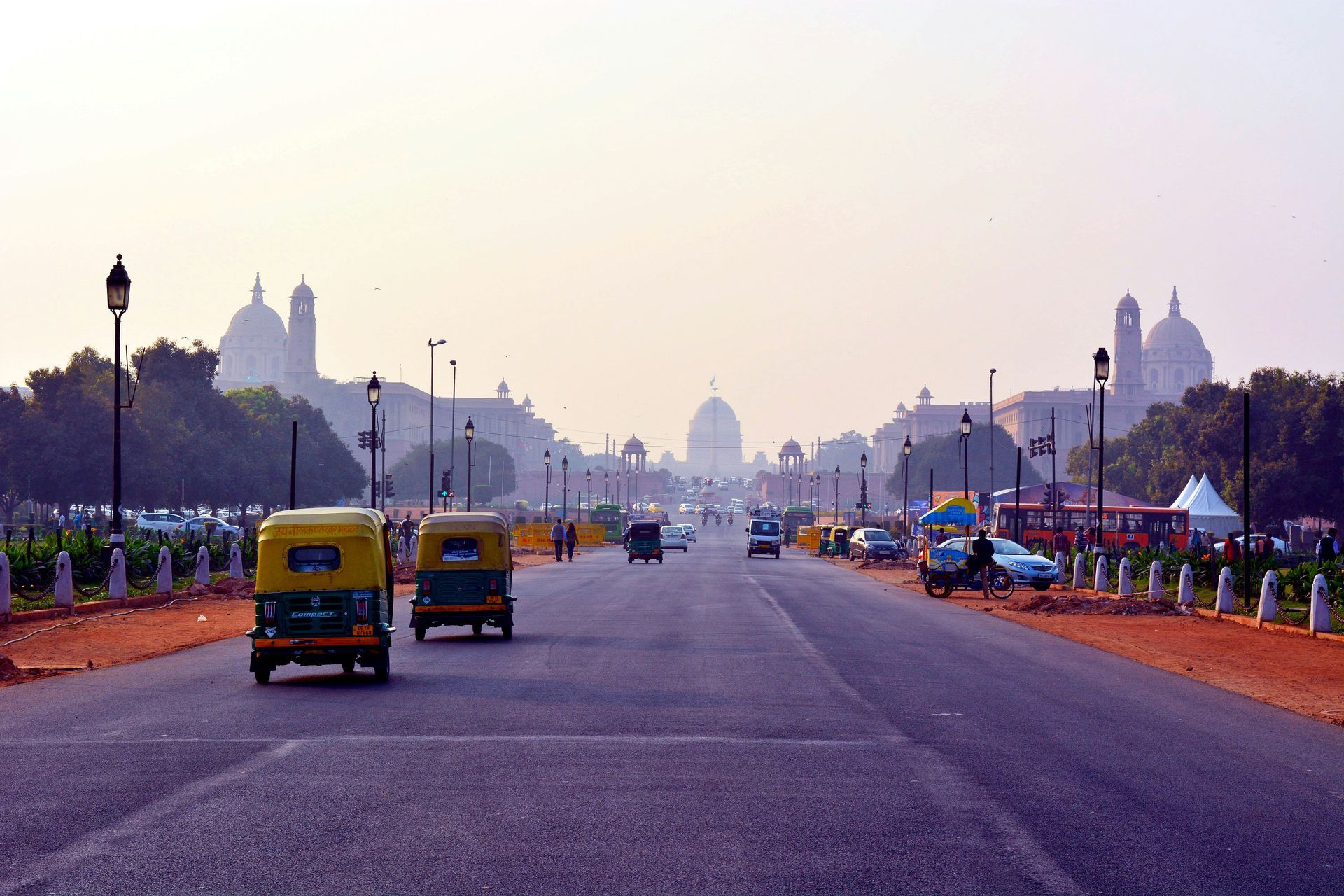 Wide avenue in Delhi with auto-rickshaws, government buildings in the distance, and a hazy sky.