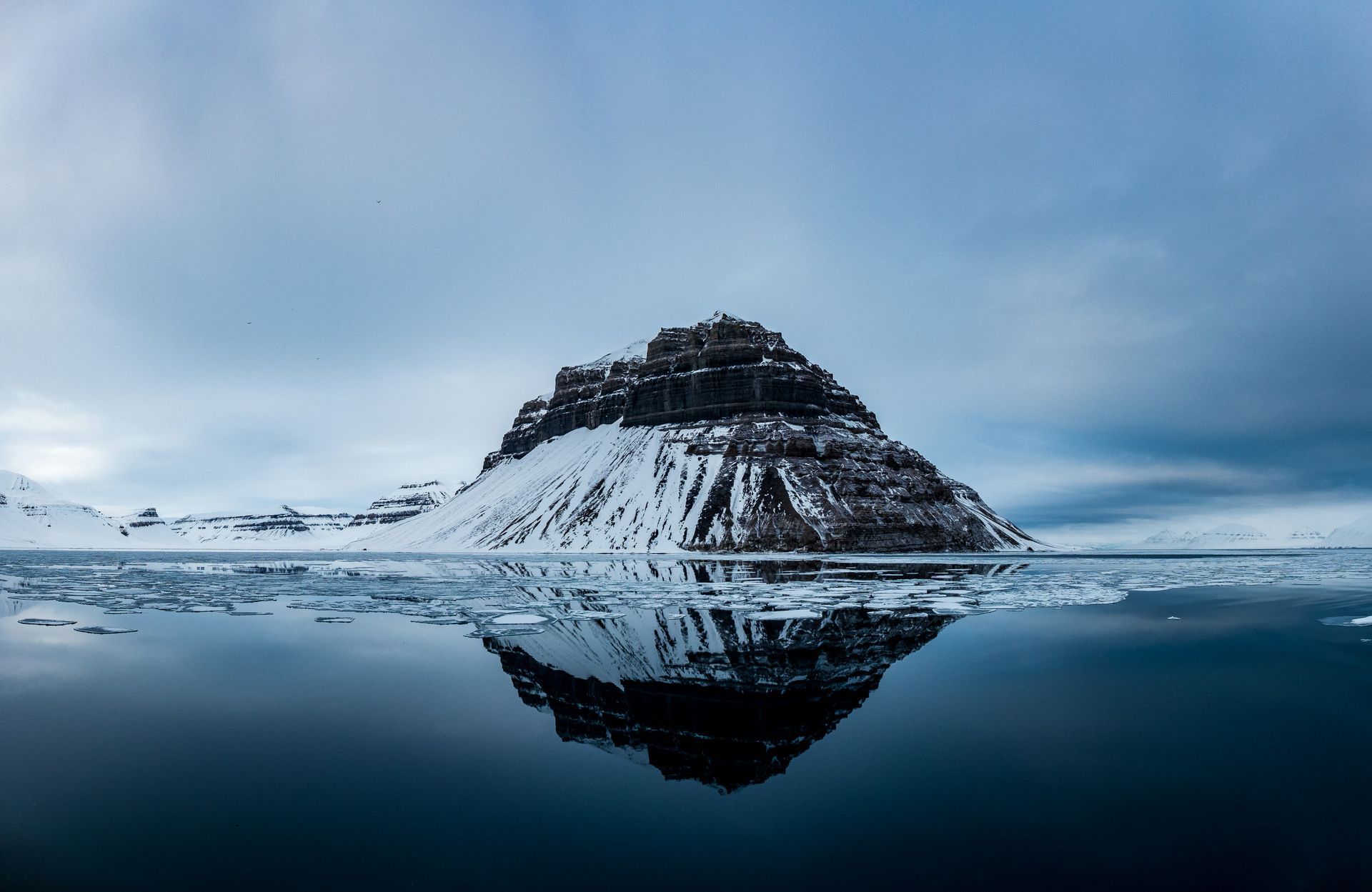 Snow-covered mountain reflected in calm, icy water under a cloudy sky in Svalbard, Norway.