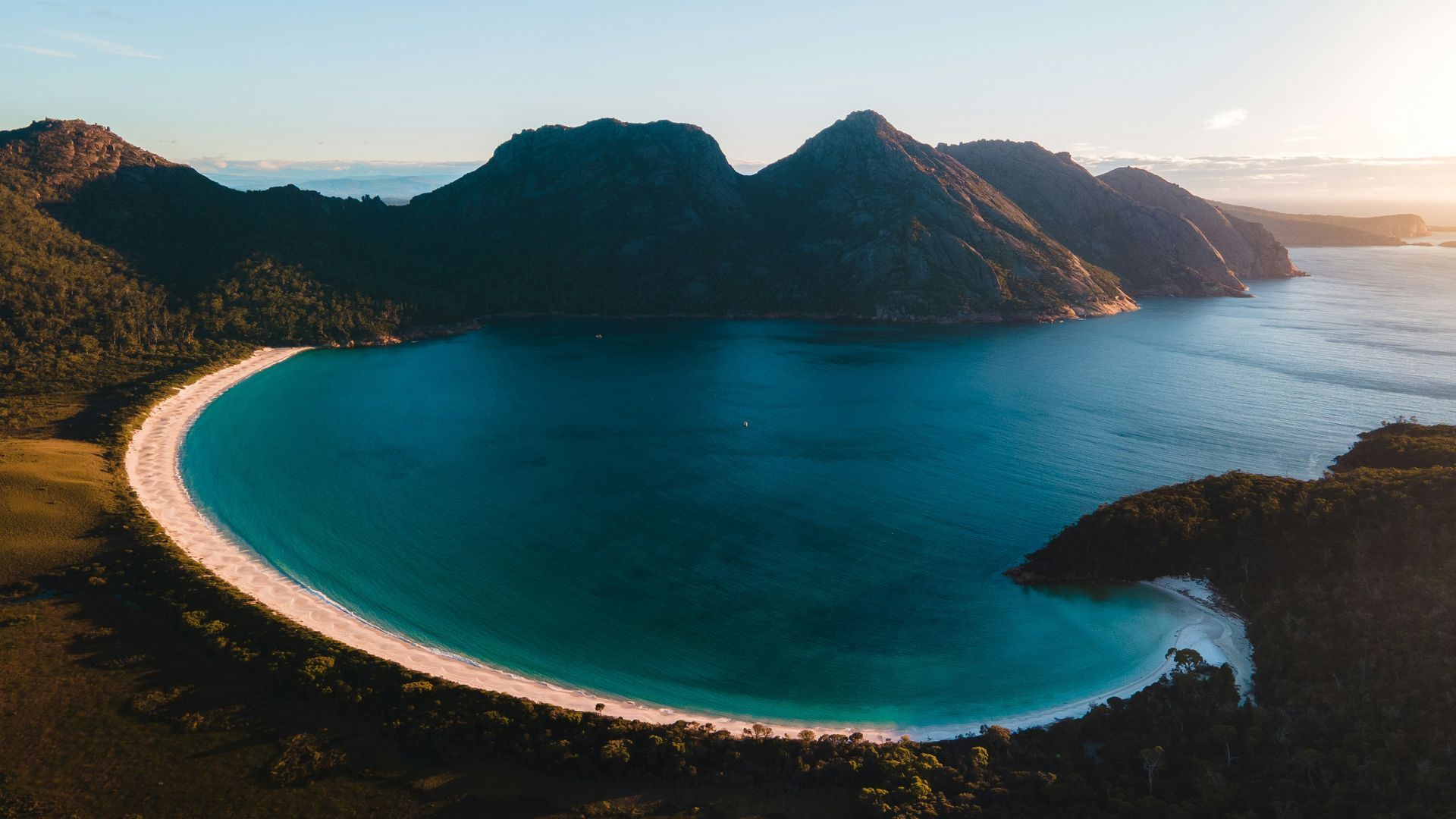 An aerial view of a circular beach in the middle of a large body of water surrounded by mountains in Tasmania, Australia.