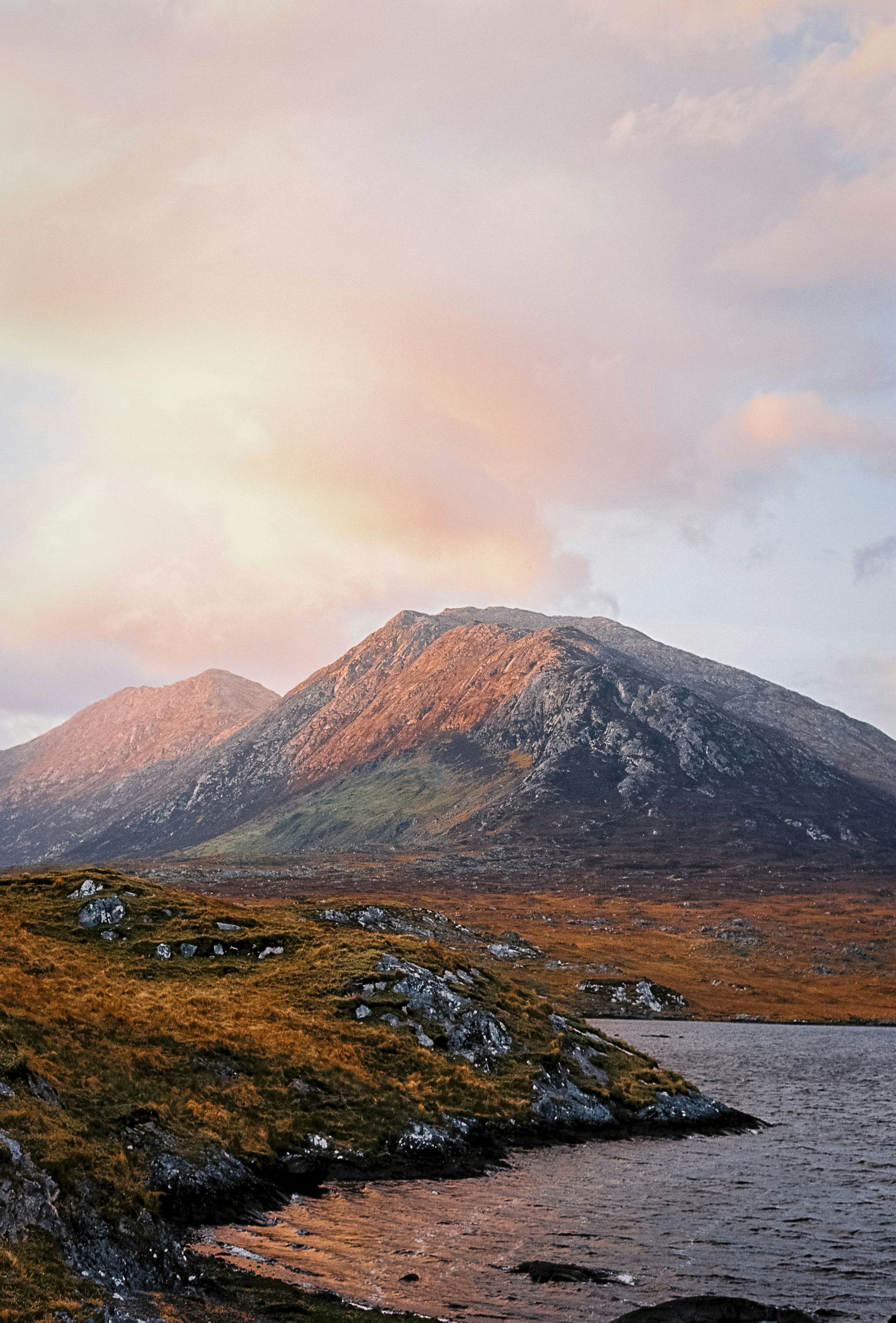 Mountains at sunset, gold and lavender sky over a lake, rocky foreground in Connemara, Ireland.