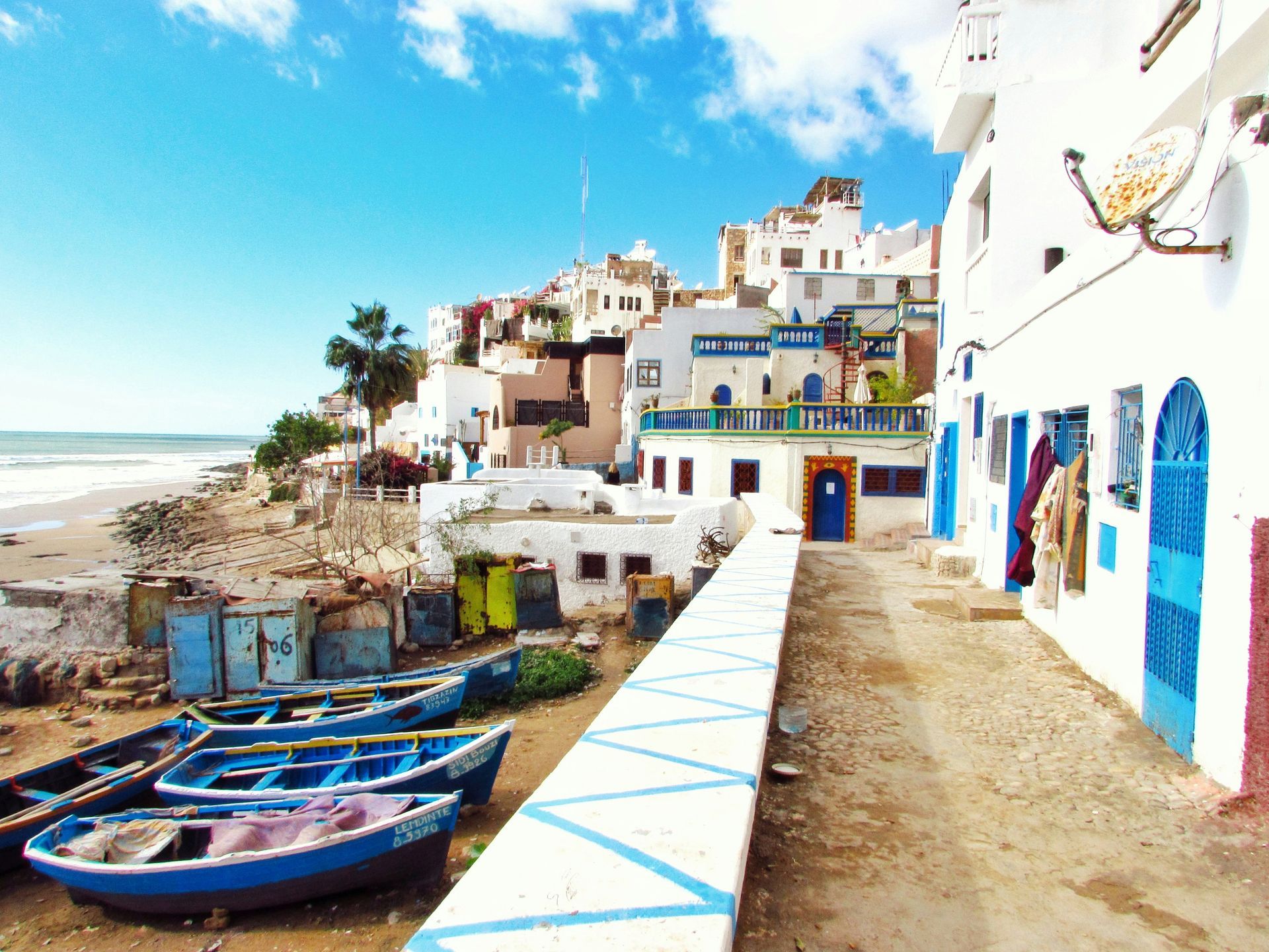 A row of boats are parked on a beach next to a white building in Taghazout, Morocco.
