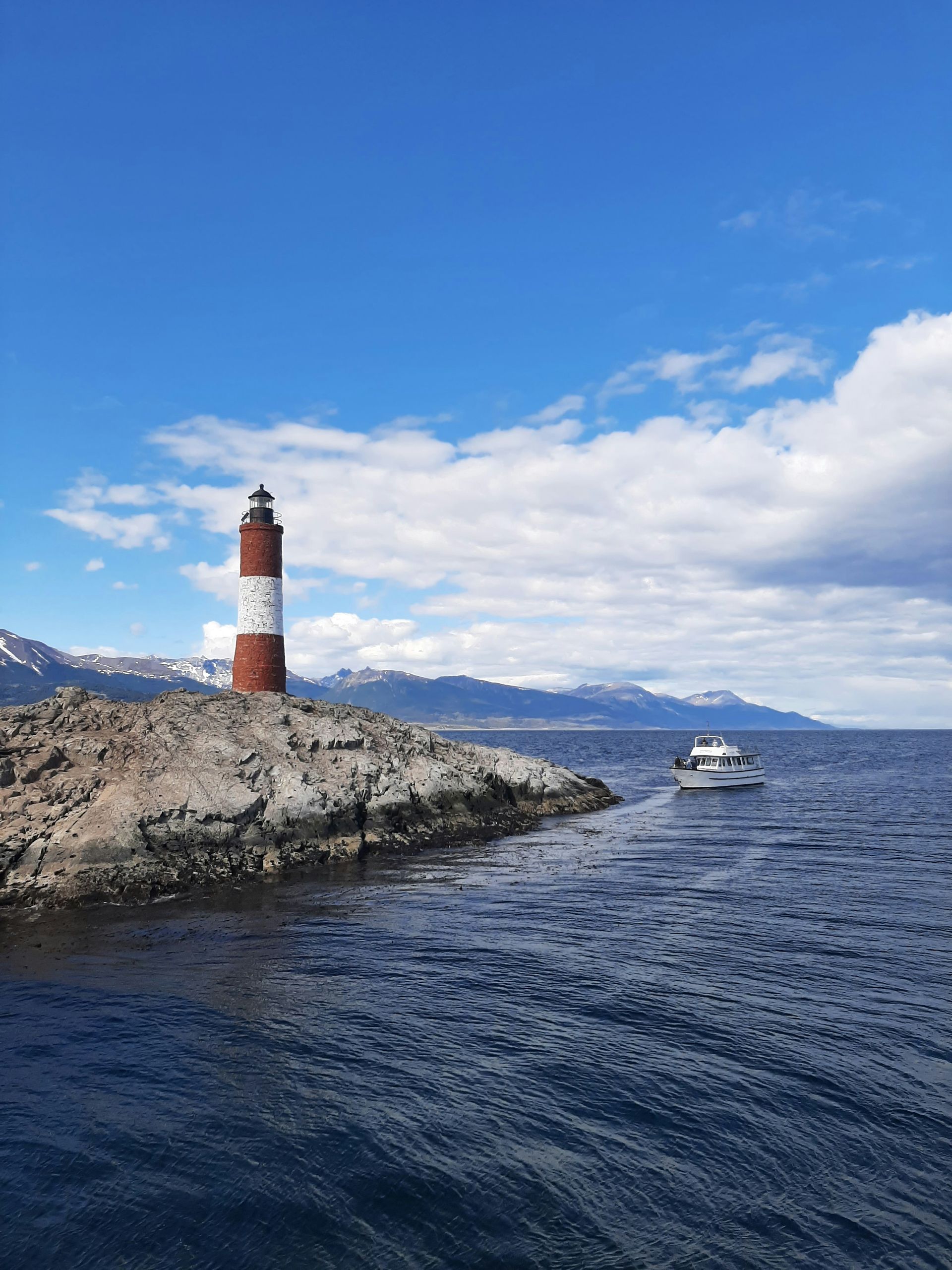 Red and white striped lighthouse on rocky island; boat approaching, mountains in background in Ushuaia, Argentina.
