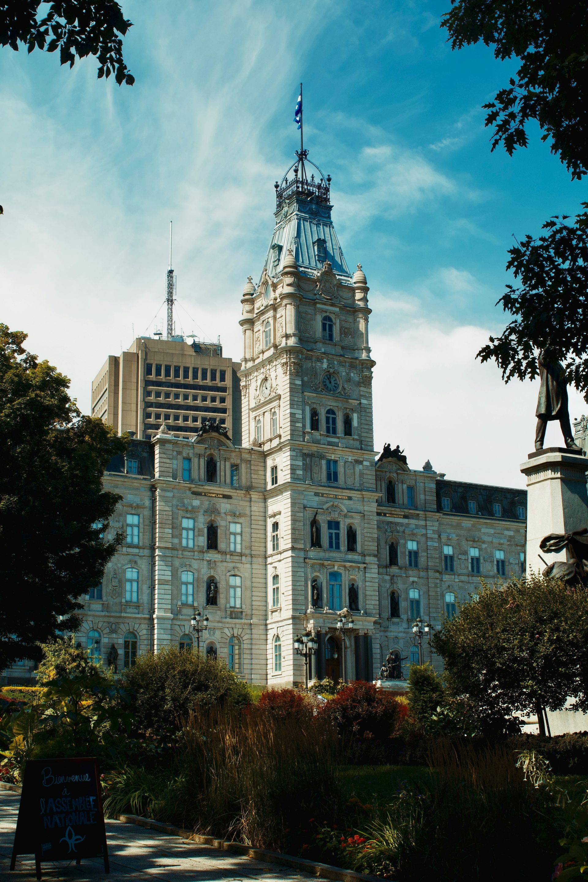 Parliament Building, Quebec City, Canada, viewed from a park with trees and a statue; the ornate stone building has a tall tower and a blue sky background.