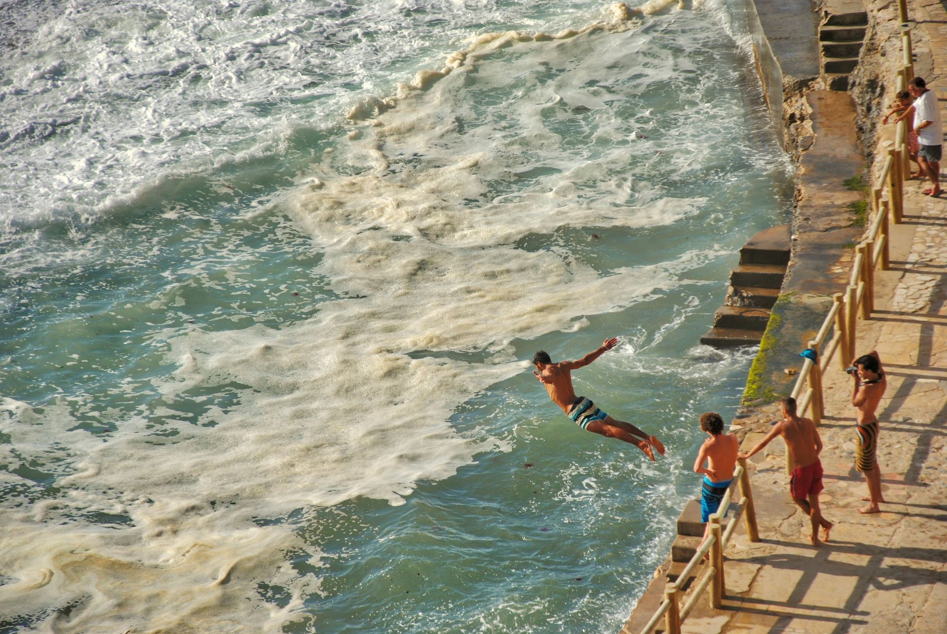 Man diving into ocean waves, people watching from a concrete ledge in Azenhas do Mar, Portugal.