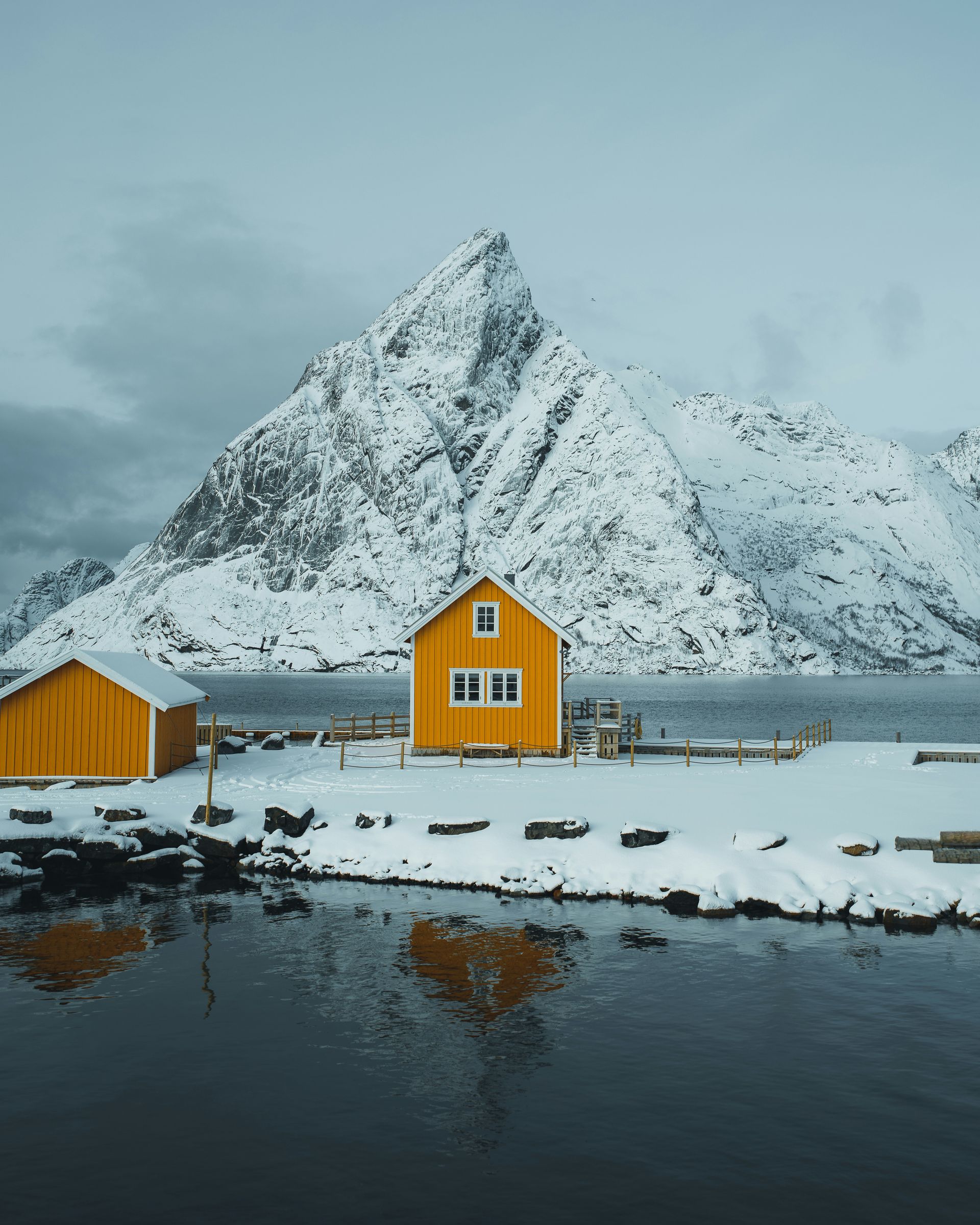 Yellow houses on snowy shore, mountain backdrop, overcast sky in Lofoten, Norway.