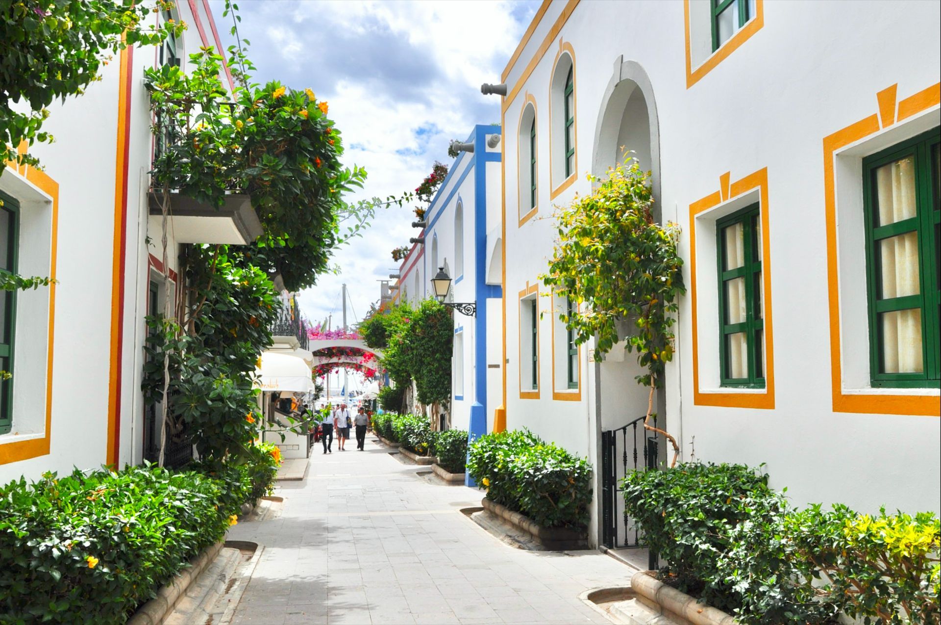 Narrow street lined with white buildings with yellow and green accents, lush greenery, and people walking in Marbella, Spain.