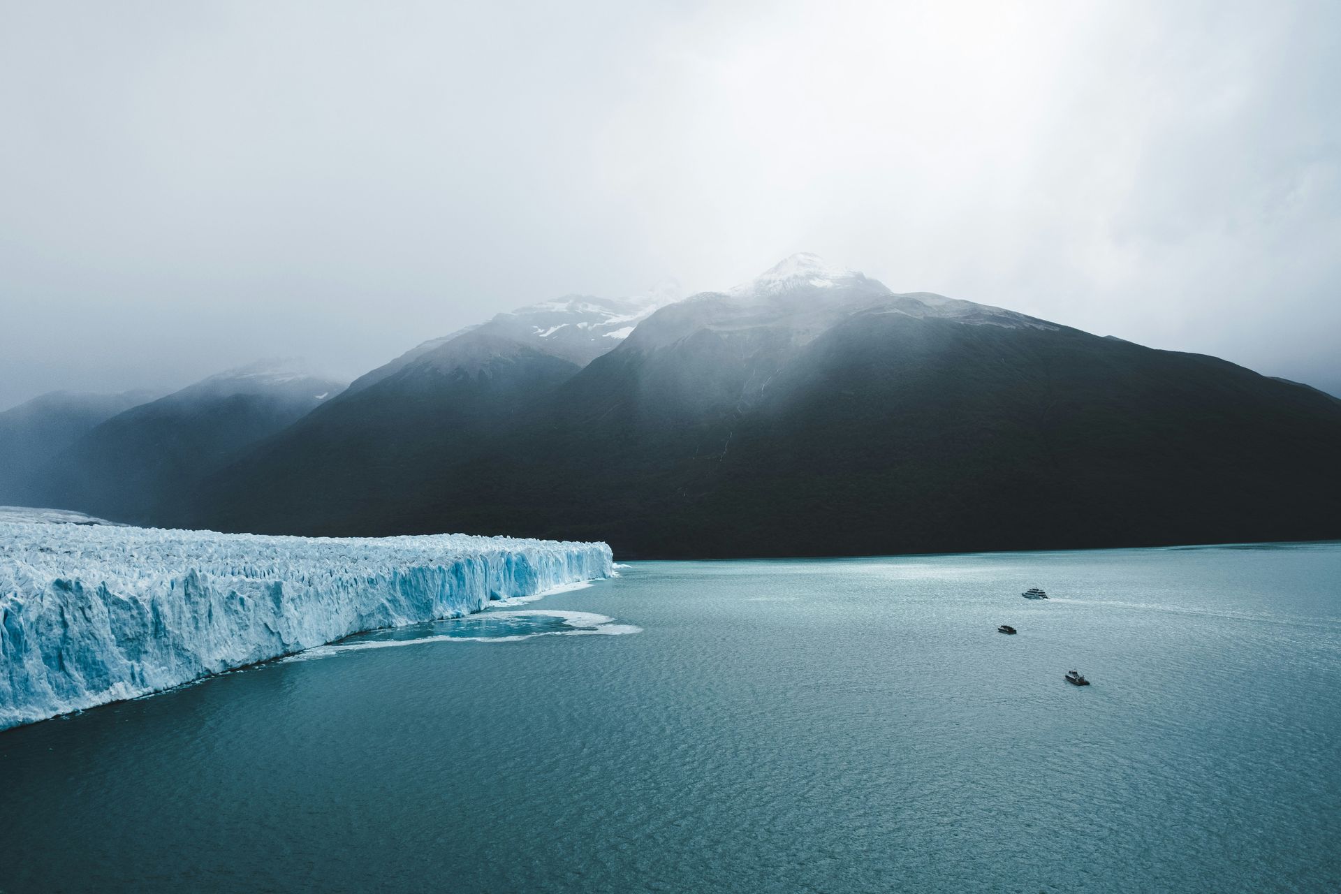 Glacier on a foggy lake with boats, dark mountains in background at Parque Nacional Los Glaciares in El Calafate, Argentina.
