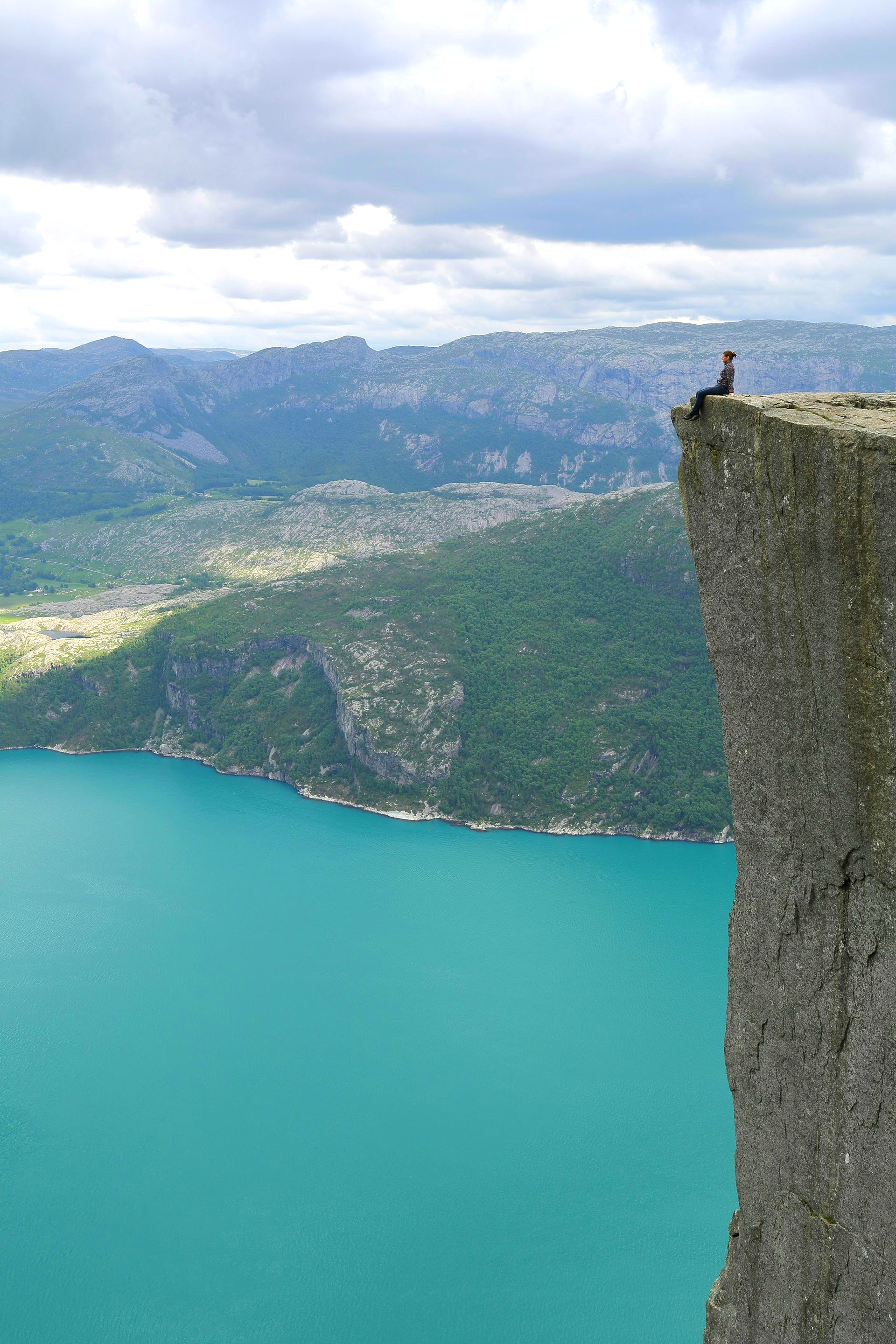 A person sitting on the cliff edge of  Pulpit Rock, overlooking the turquoise Lysefjord and green mountains.