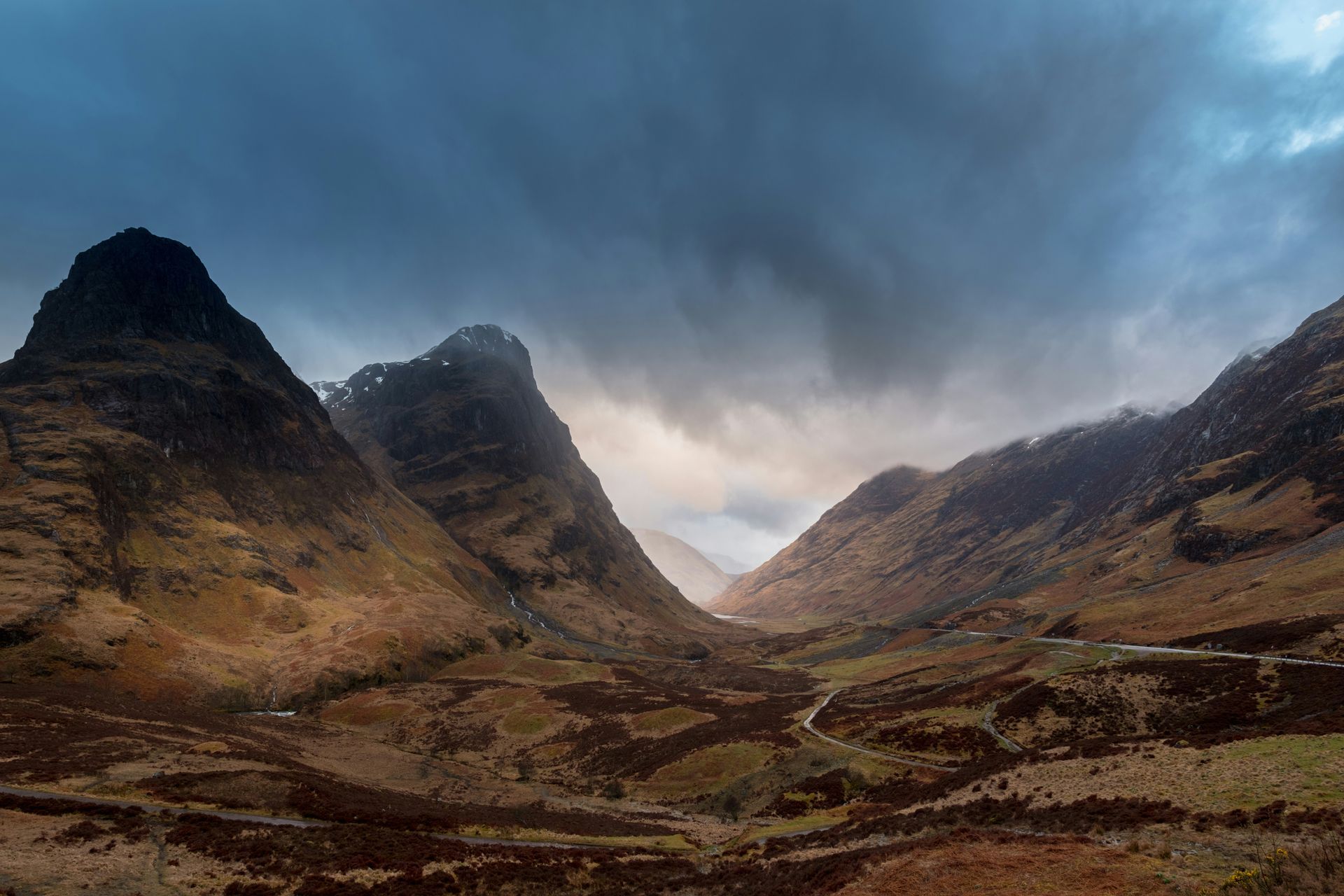 Mountains in a valley under a dramatic, cloudy sky; brown landscape with small road in Glencoe, Scotland.