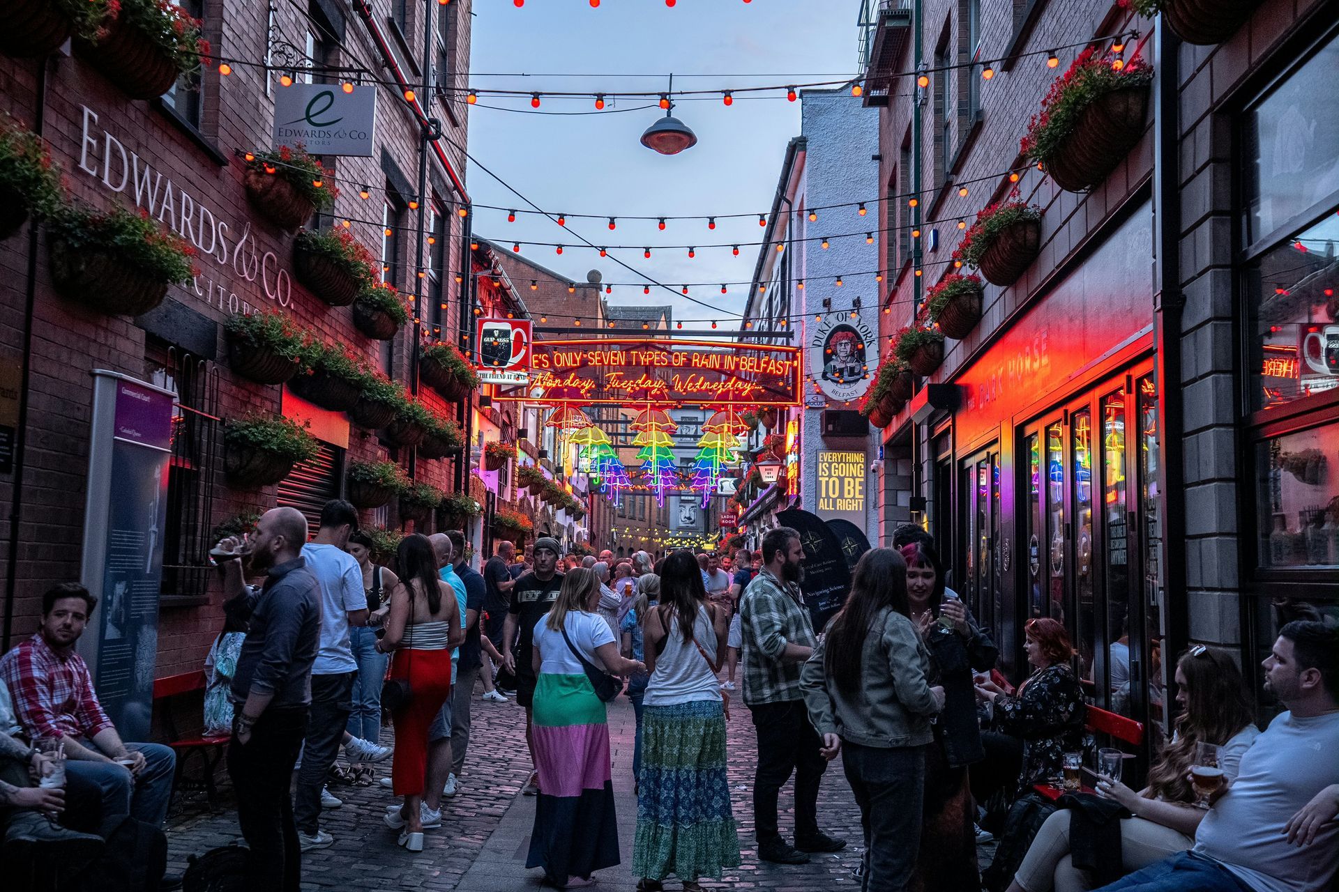 A crowded street lined with bars, decorated with colorful lights, where people socialize in Belfast, Northern Ireland.