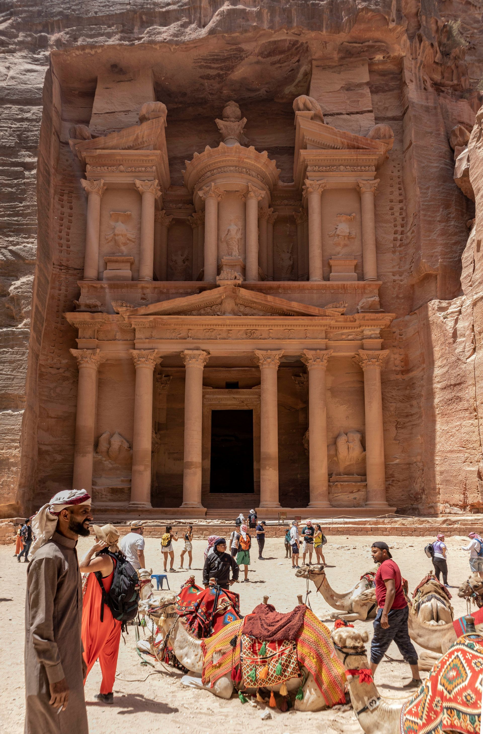 The Treasury in Petra, Jordan, carved into sandstone cliffs. Tourists and camels in foreground.