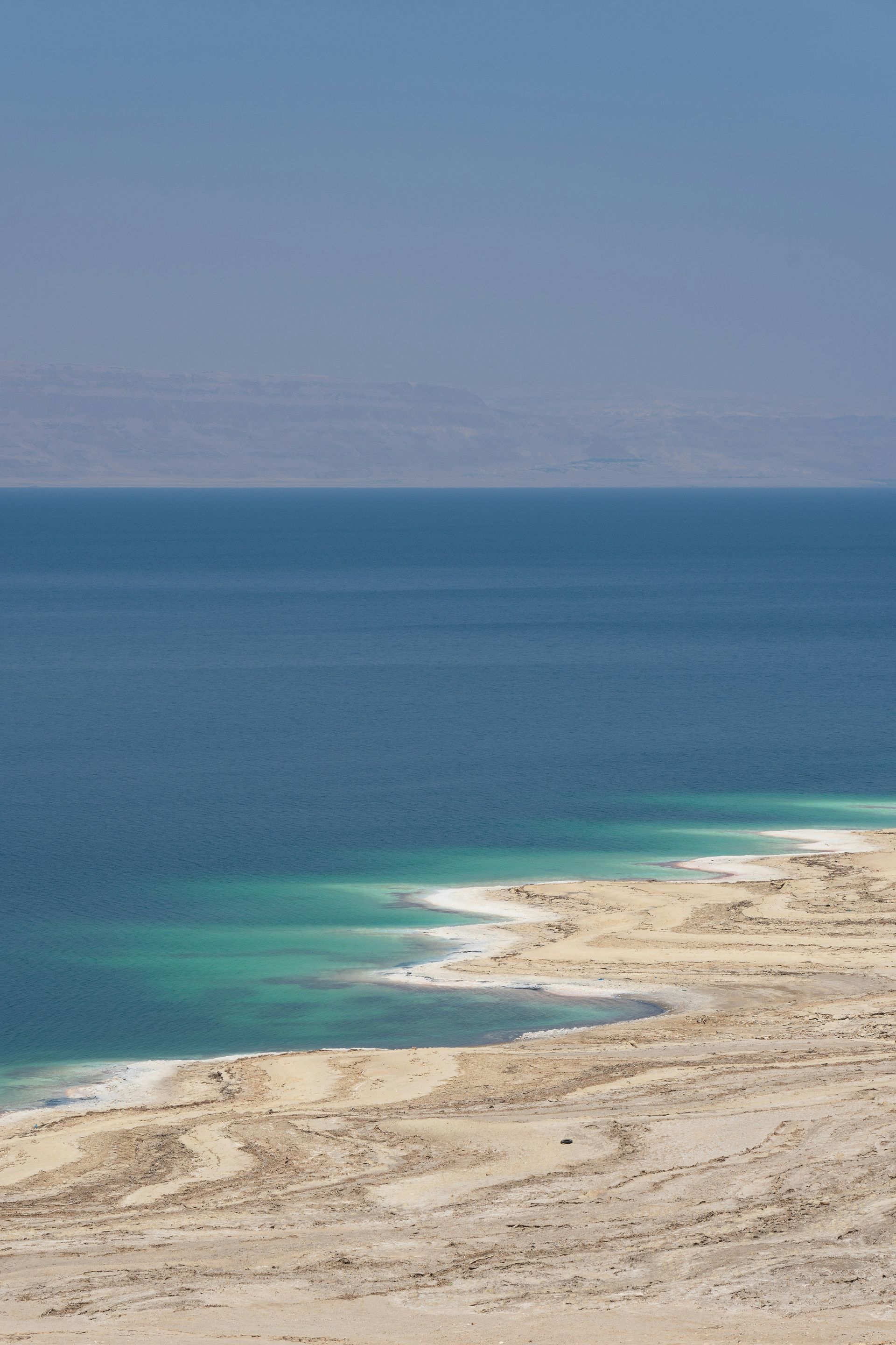 Dead Sea shoreline with blue water meeting a light-colored beach. Distant mountains in haze in Jordan.