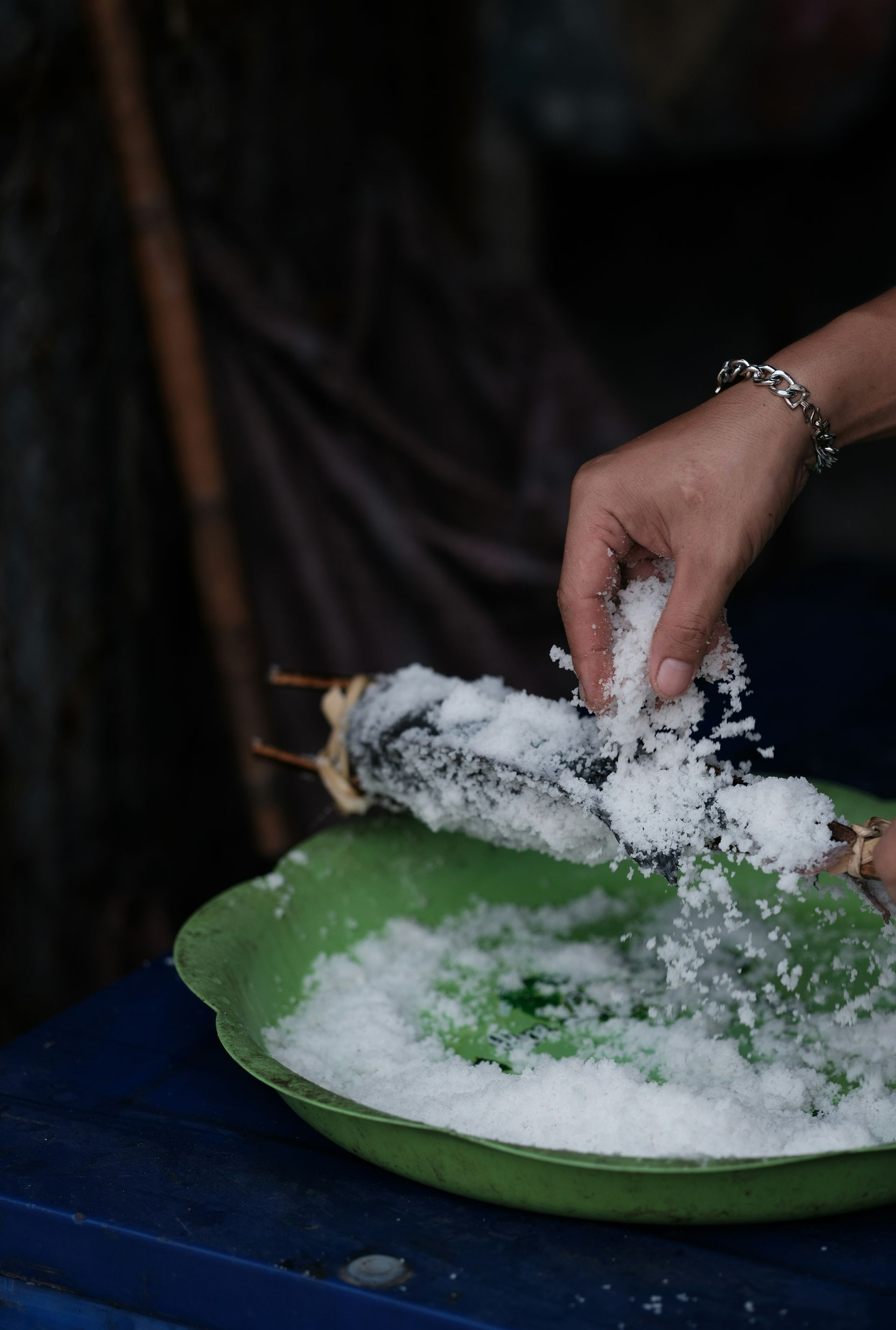 A person sprinkles salt onto fish held on a bamboo skewer over a green bowl in Laos.