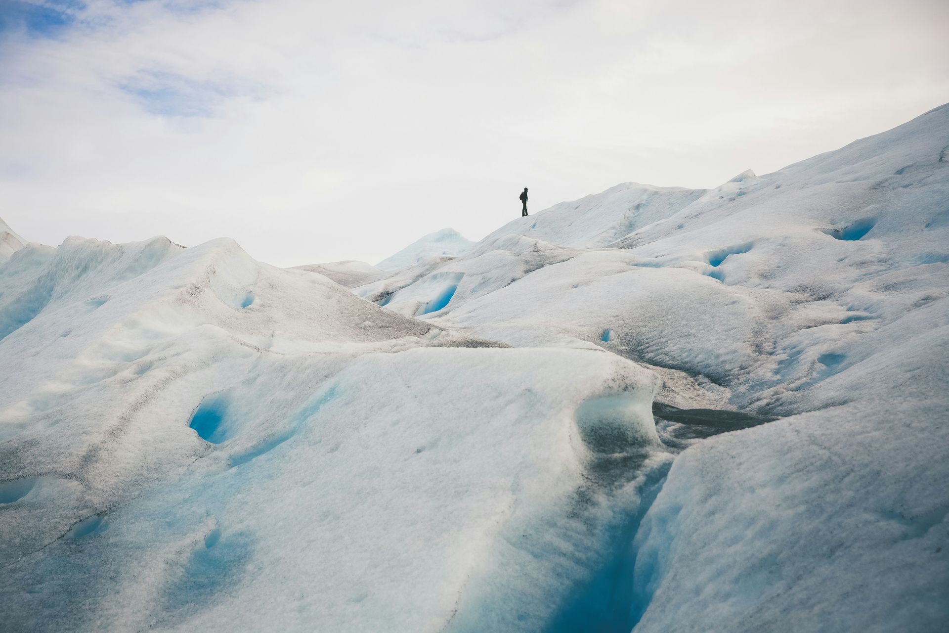 Person on glacier, vast icy terrain with blue pools, overcast sky at Parque Nacional Los Glaciares in El Calafate, Argentina.