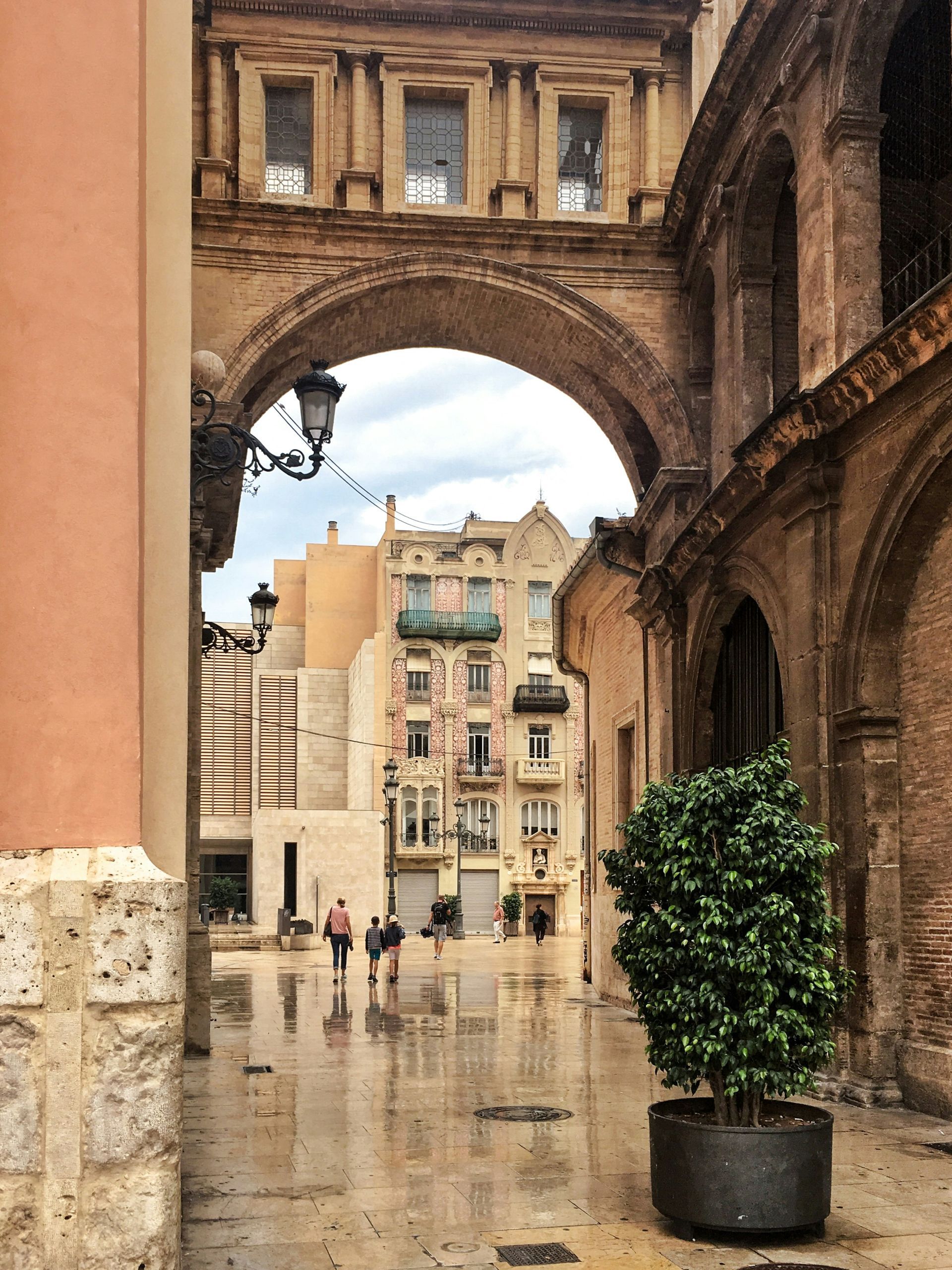 View through archway: People walking on wet cobblestones towards buildings, Valencia, Spain.