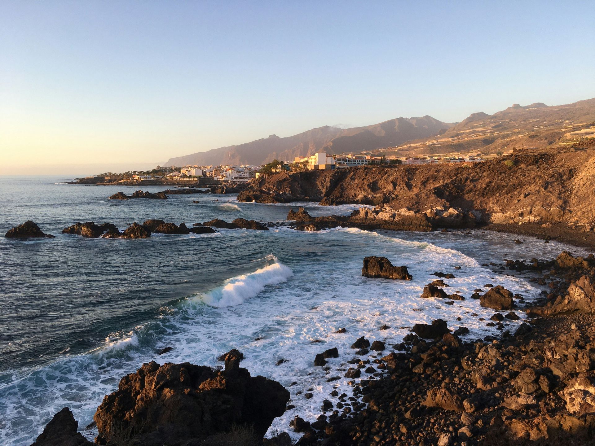 Rocky coastline with waves crashing, a town, and mountains in the background on the Canary Islands, Spain.