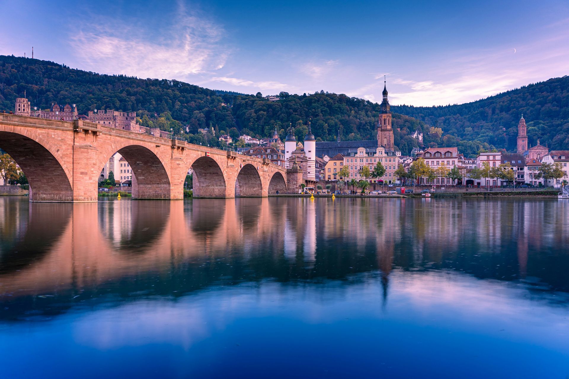 Stone bridge over calm water, Heidelberg, Germany, with buildings and hills in the background.