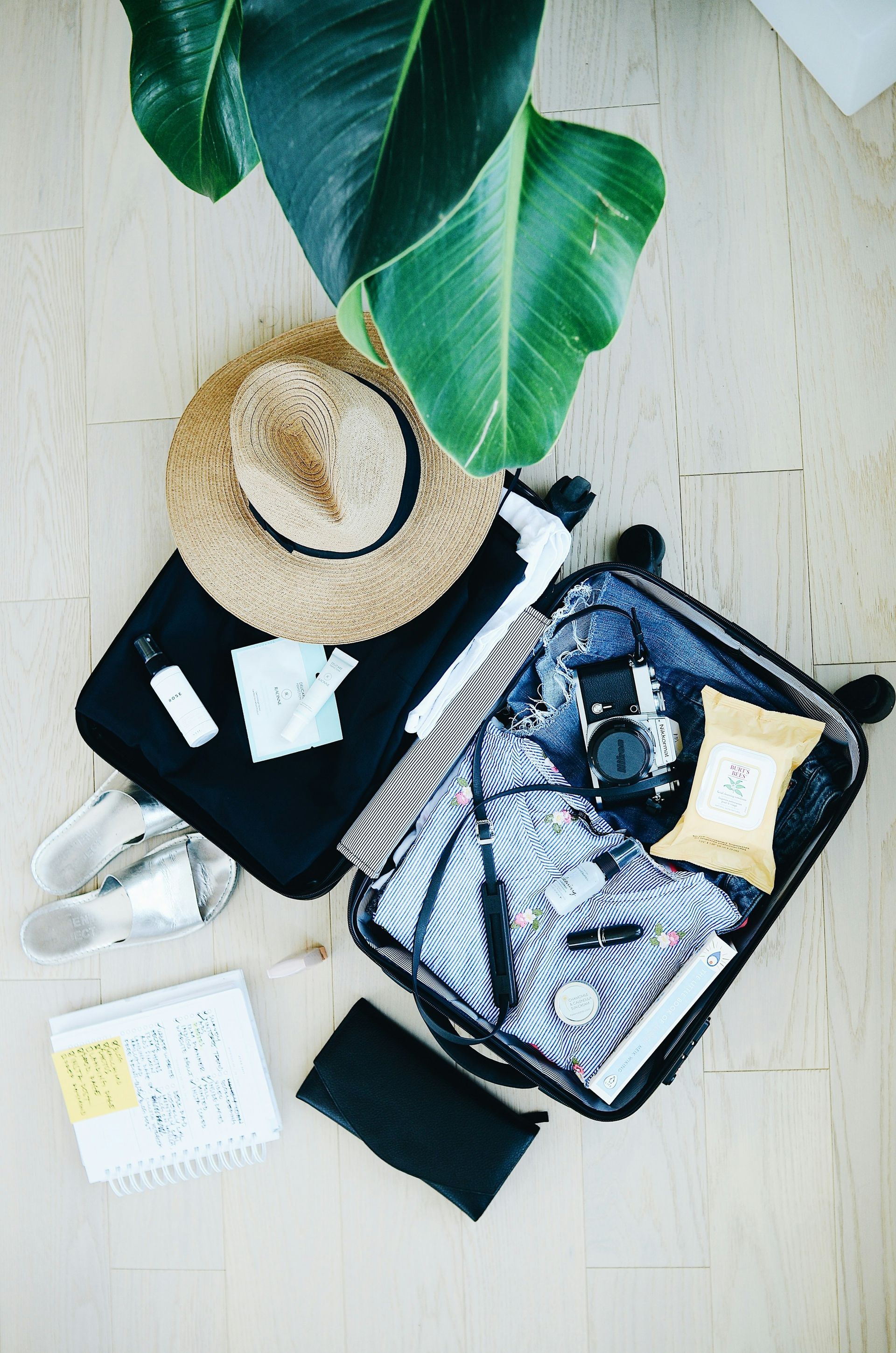 A suitcase filled with clothes and a hat is sitting on a wooden floor next to a plant.