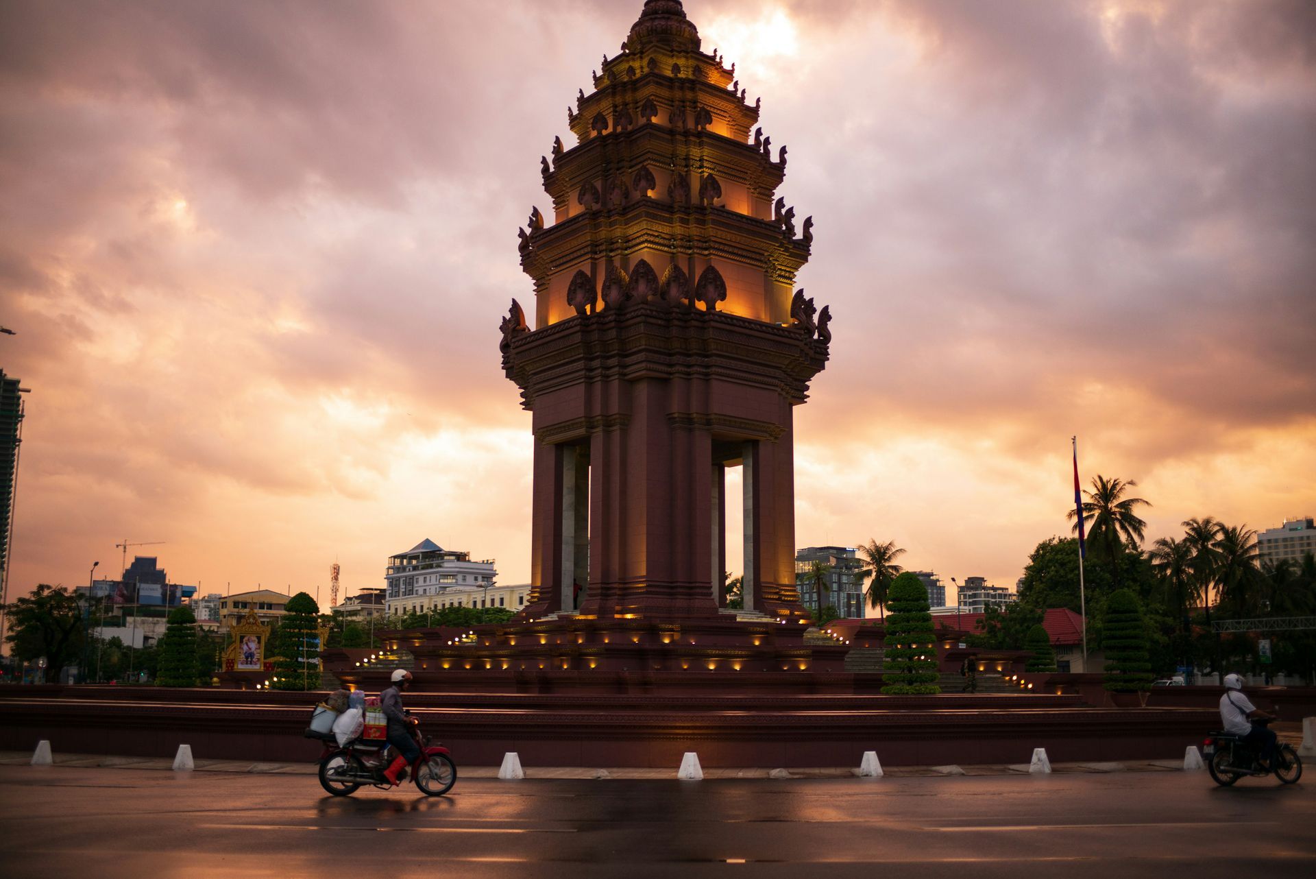 Cambodian Independence Monument at sunset with two motorcyclists passing by.