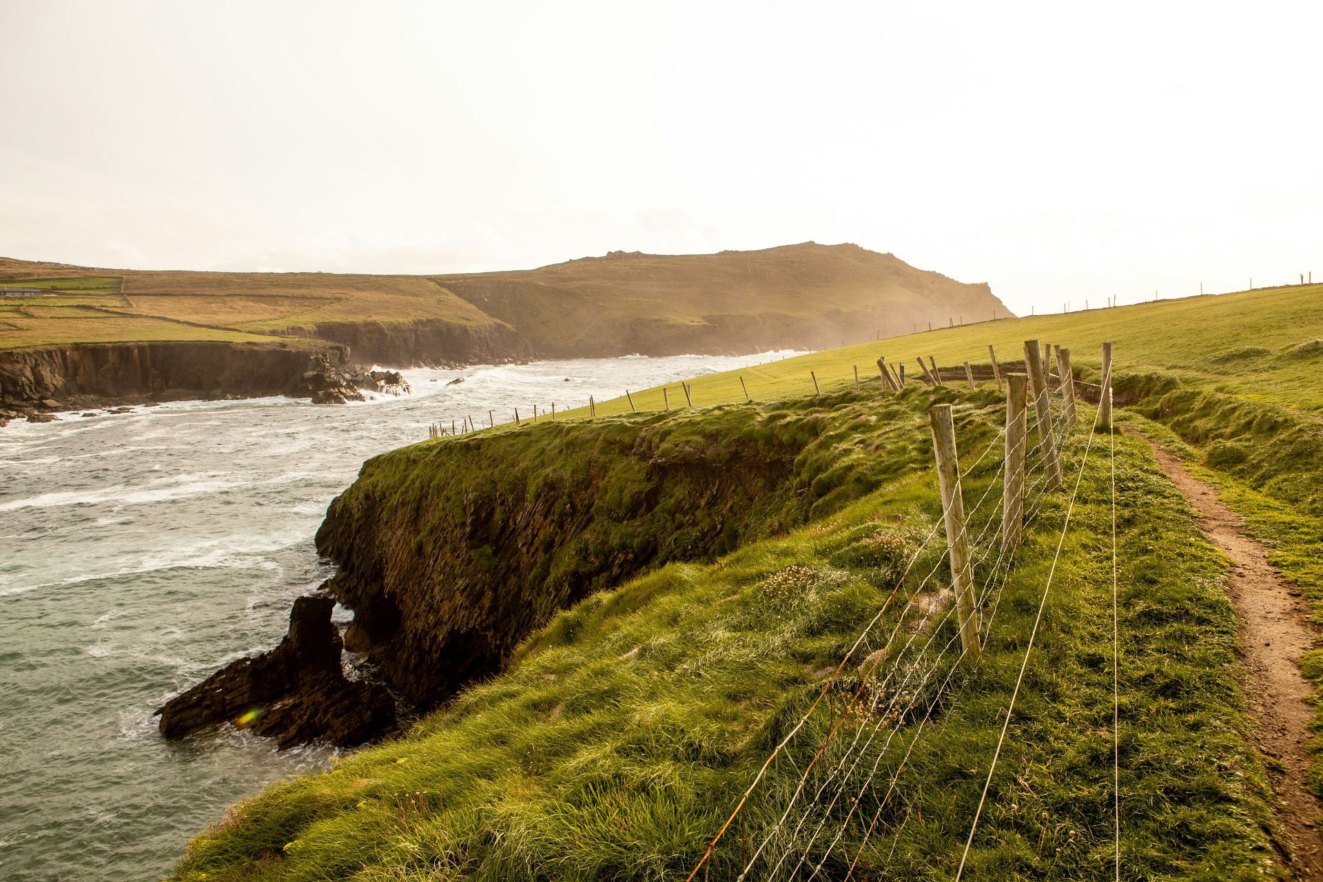 Grassy cliffside path with a fence, overlooking a choppy ocean and distant hills on the Dingle Peninsula, Ireland.