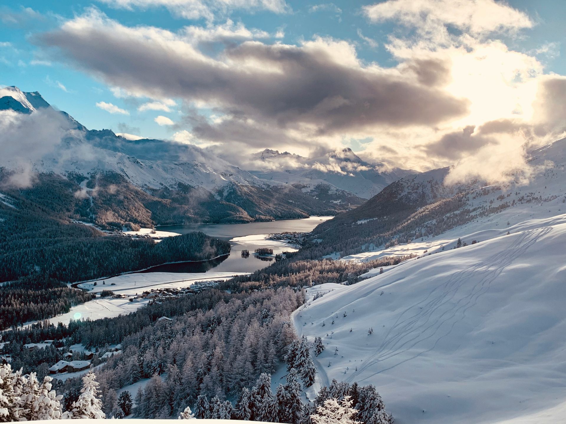 Snow-covered mountains and pine trees surround a lake; a bright sky with clouds shines overhead in St. Moritz, Switzerland. 