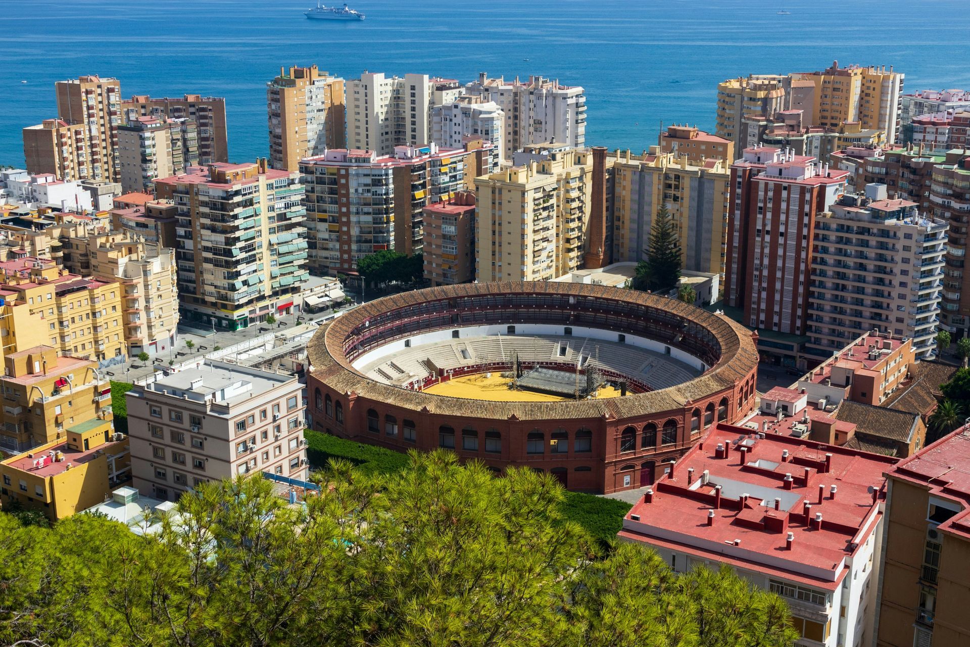 Aerial view of a bullring in Malaga, Spain, surrounded by beige buildings and the blue Mediterranean Sea.