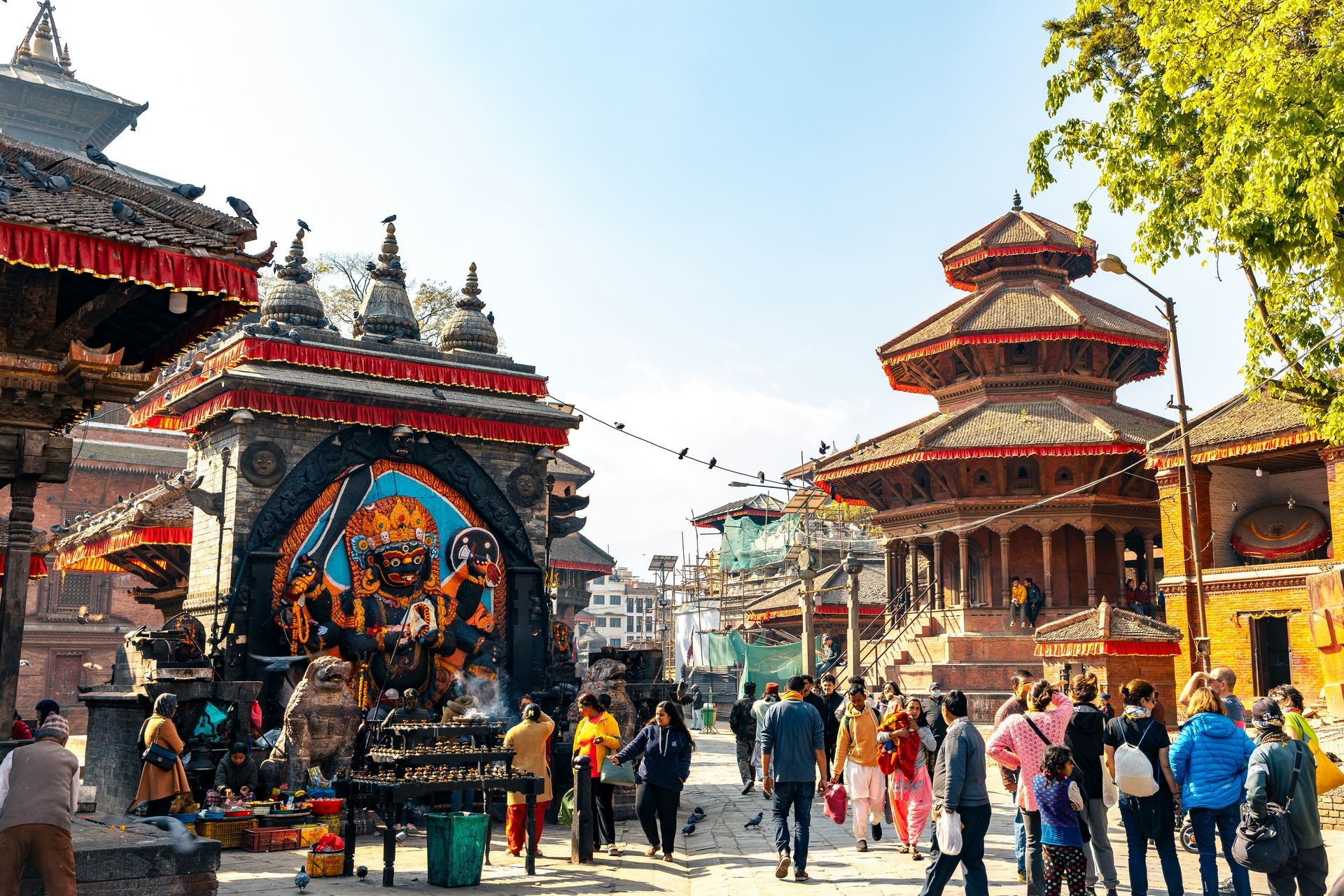 People walk in a bustling square with ornate Nepalese temples in Kathmandu, Nepal.