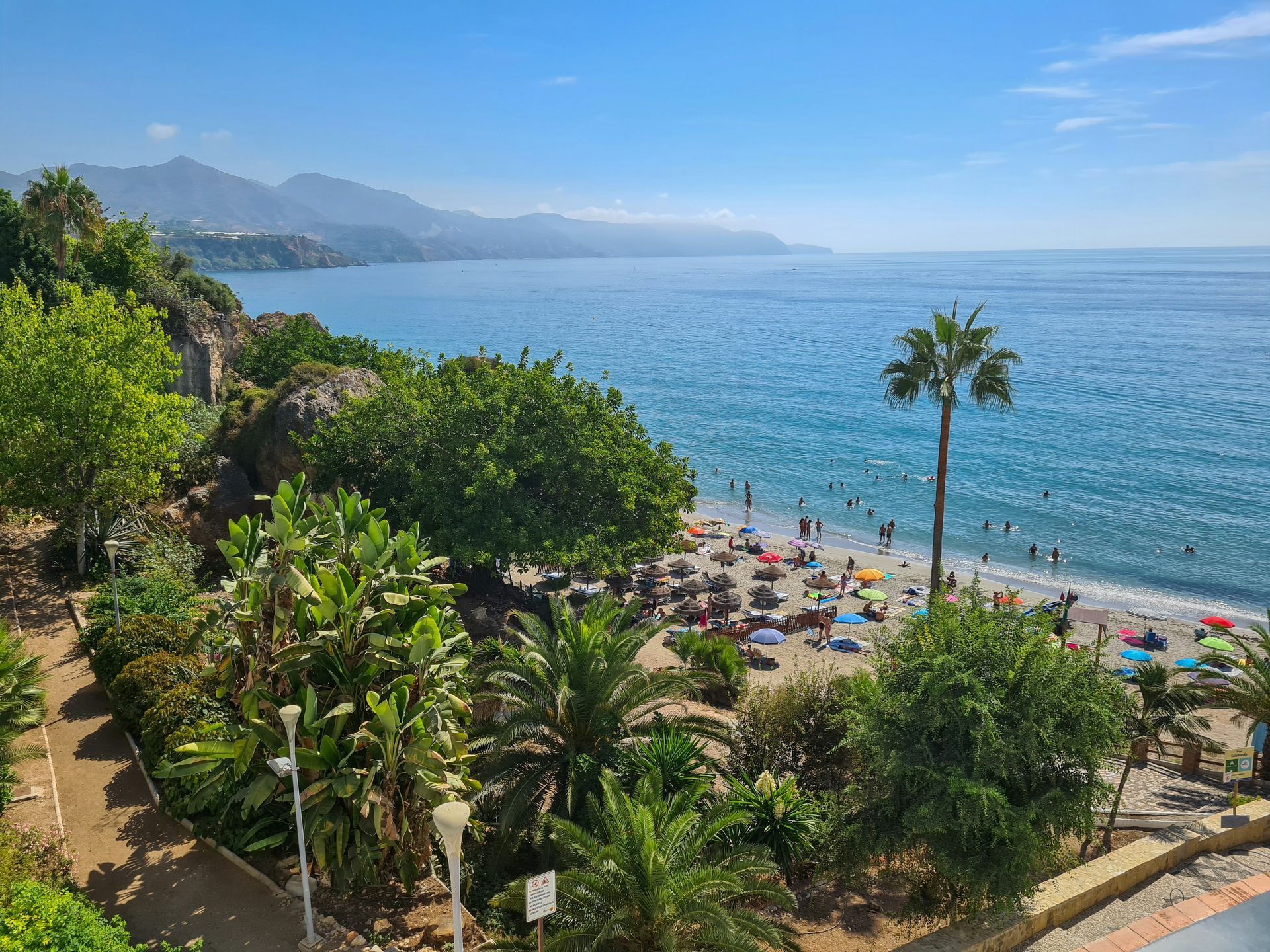 Beach scene with clear blue water, sandy shore, people, palm trees, and green vegetation in Málaga, Spain.