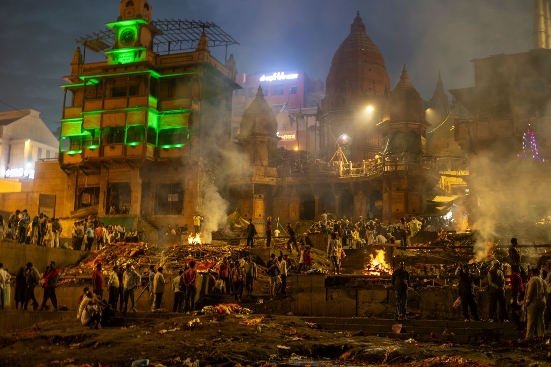 Cremation ghat in Varanasi, India, at night, with multiple funeral pyres burning, people present, smoke and buildings in the background.
