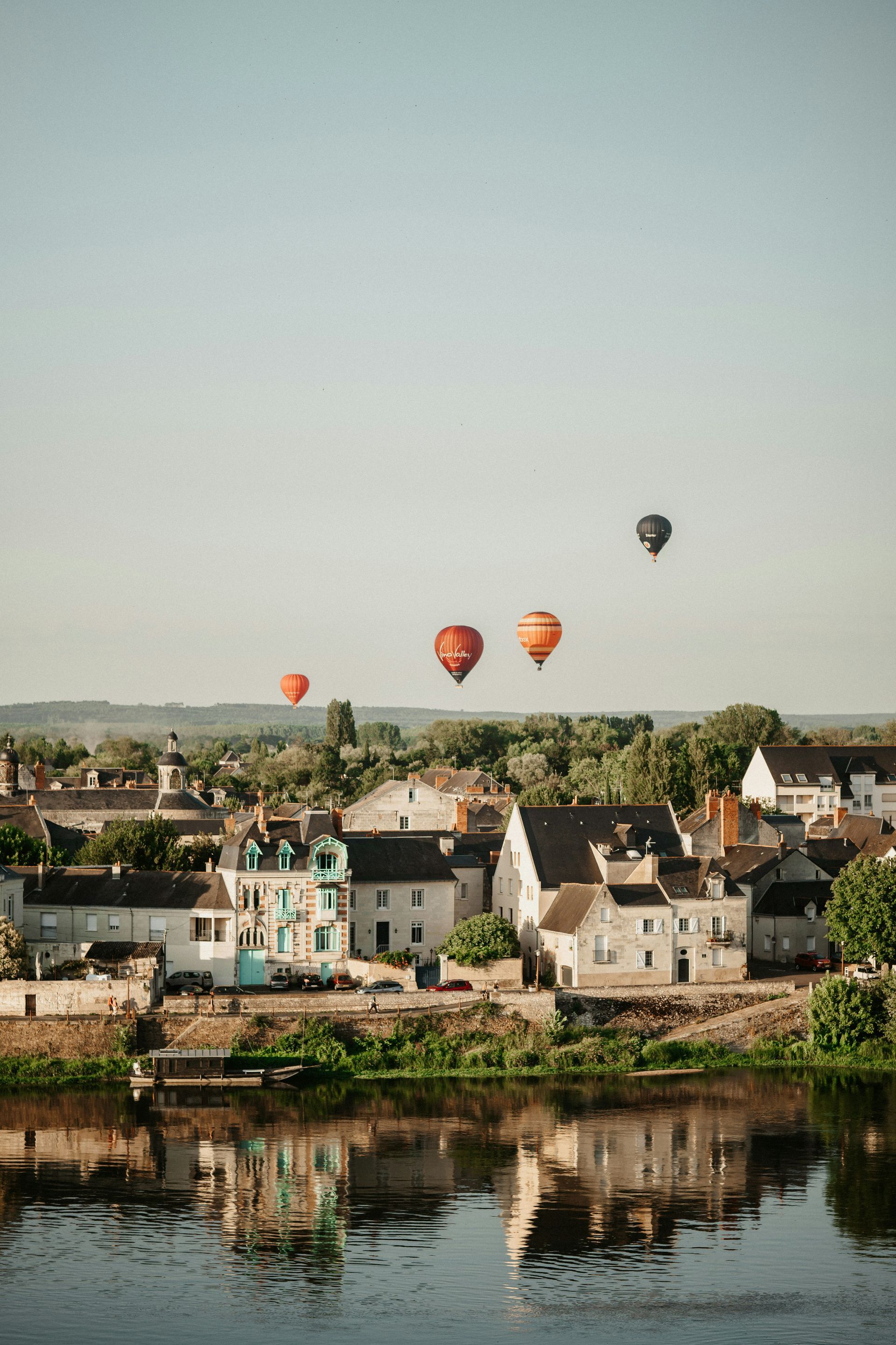 A group of hot air balloons are flying over a small town over a body of water in the Loire Valley, France.