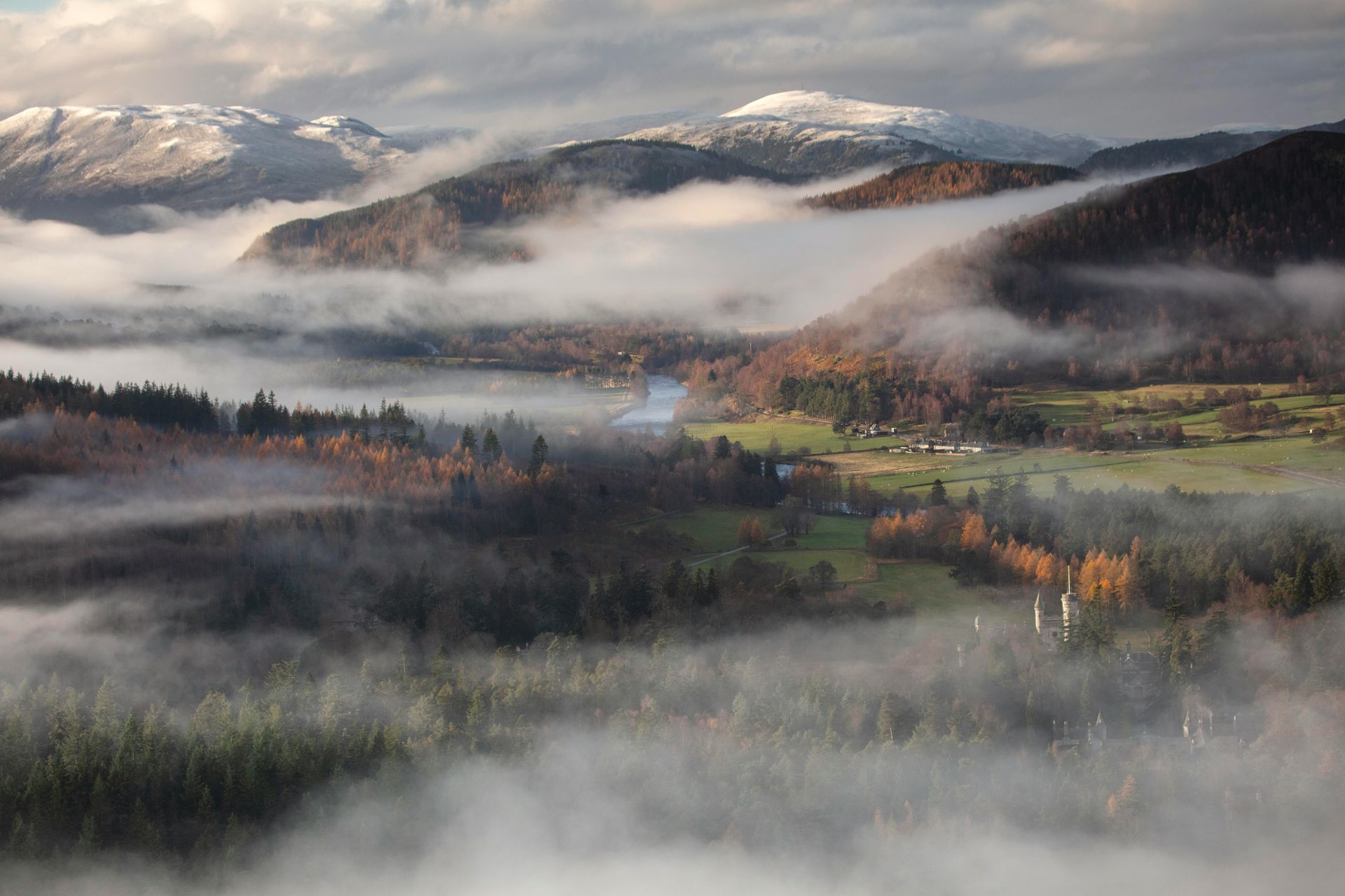 Rolling mountains partially shrouded in fog; fall foliage, green fields, and snow-capped peaks in the Cairngorms, Scotland.