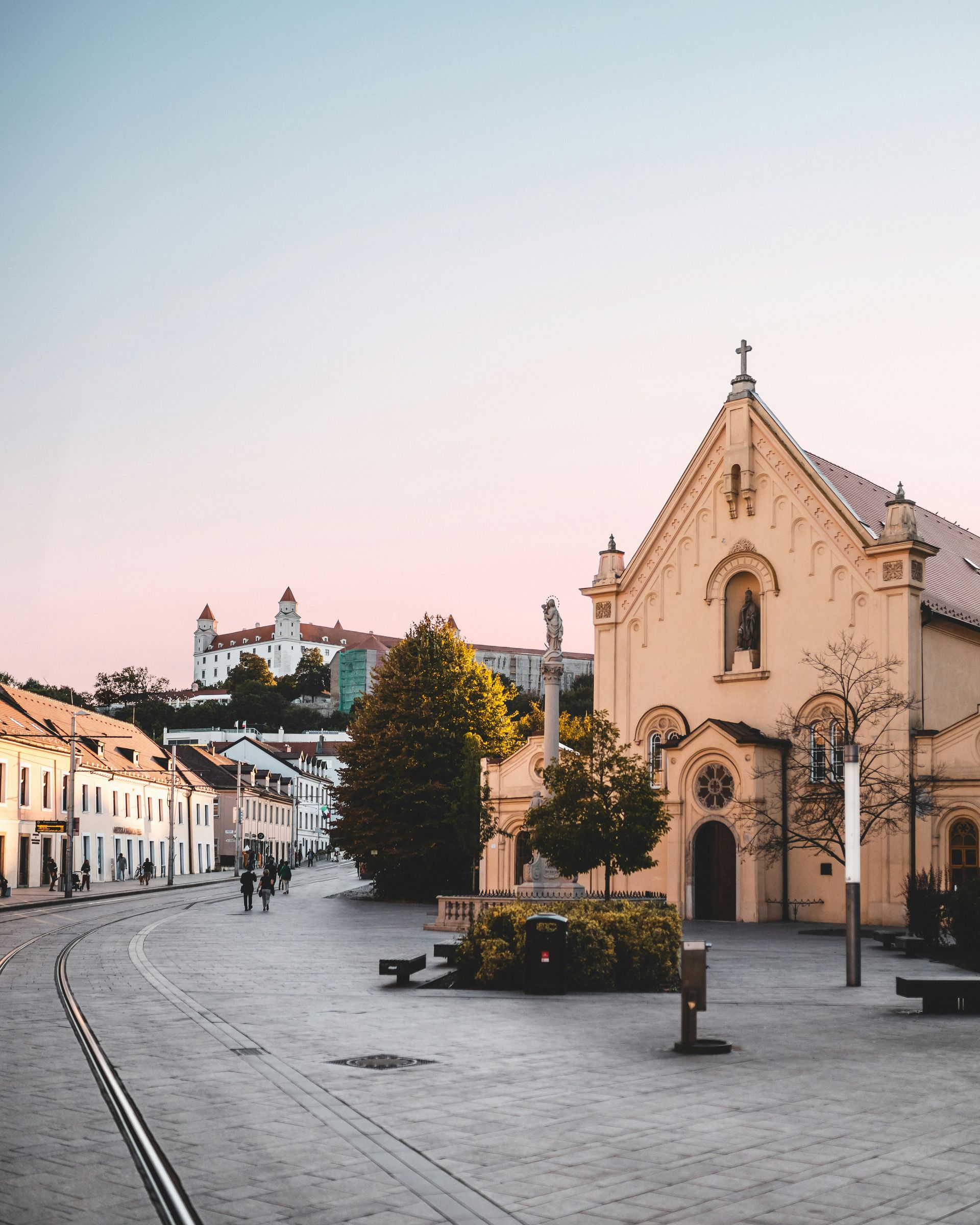 Cobblestone street with church, buildings, and castle on a hill under a pink-toned sky.