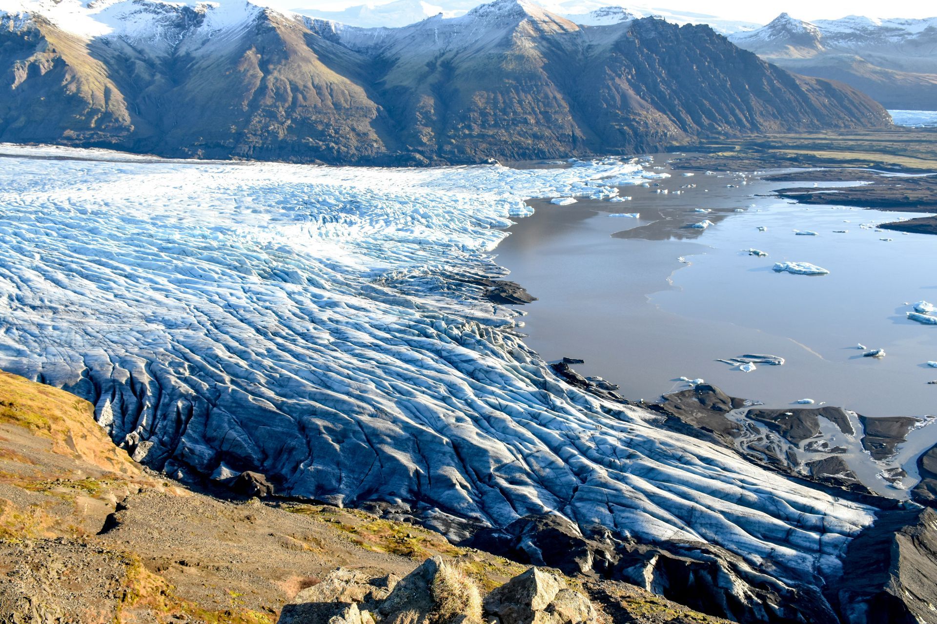Glacier flows into a lake, mountains in the background. Snowy peaks, icy blue and brown water at Vatnajökull National Park, Iceland.