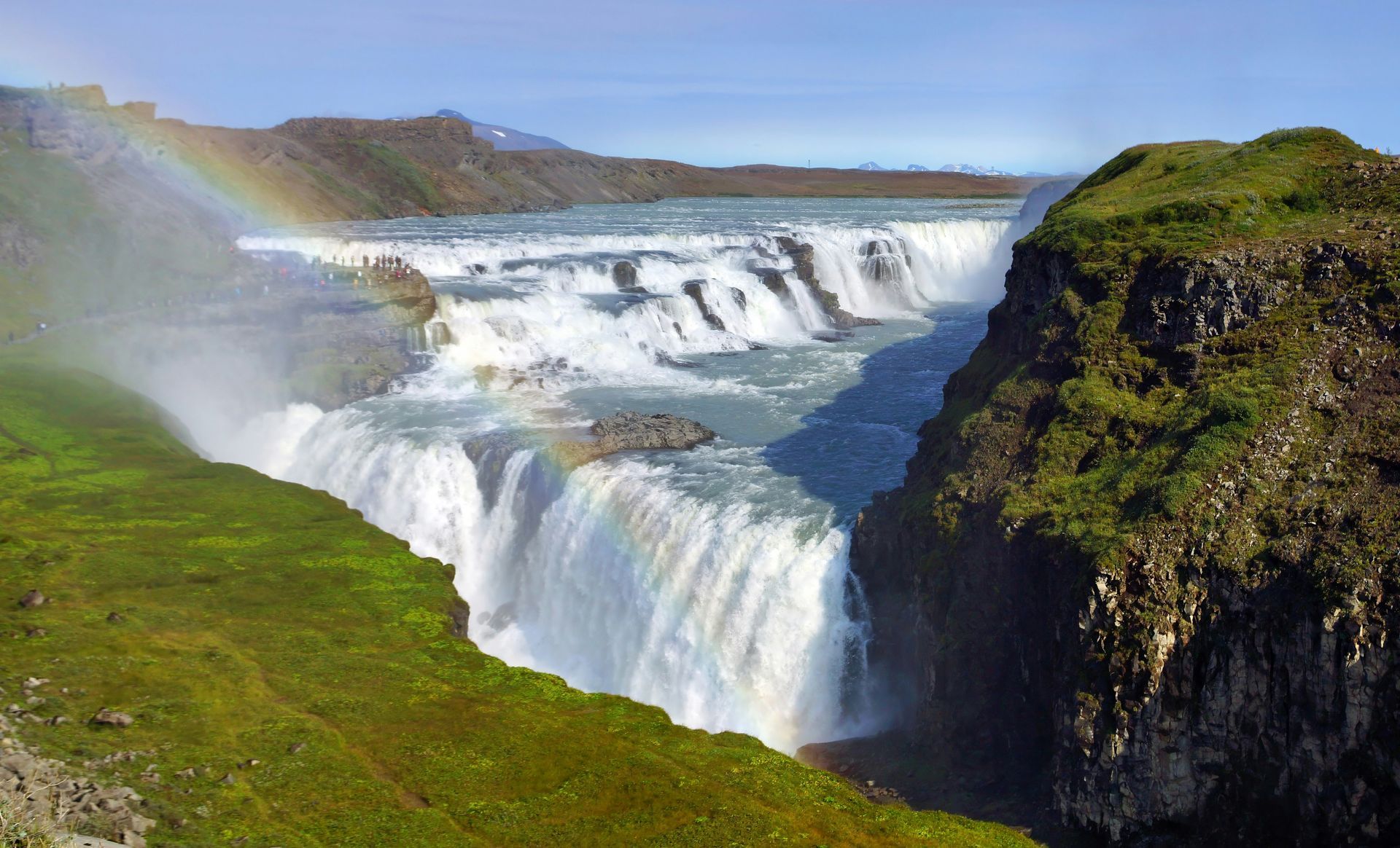Gullfoss Falls cascading into a gorge; rainbow arcs across the spray. Green cliffs and blue sky in Iceland.